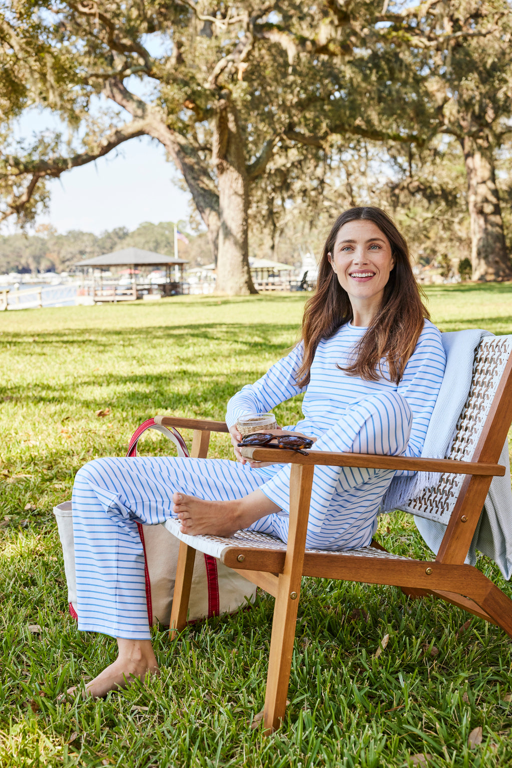 A woman wearing the Pima Crew Long-Long Set in Baltic Blue sits smiling in a wooden chair on a grassy lawn, with trees and a lake in the sunny background.