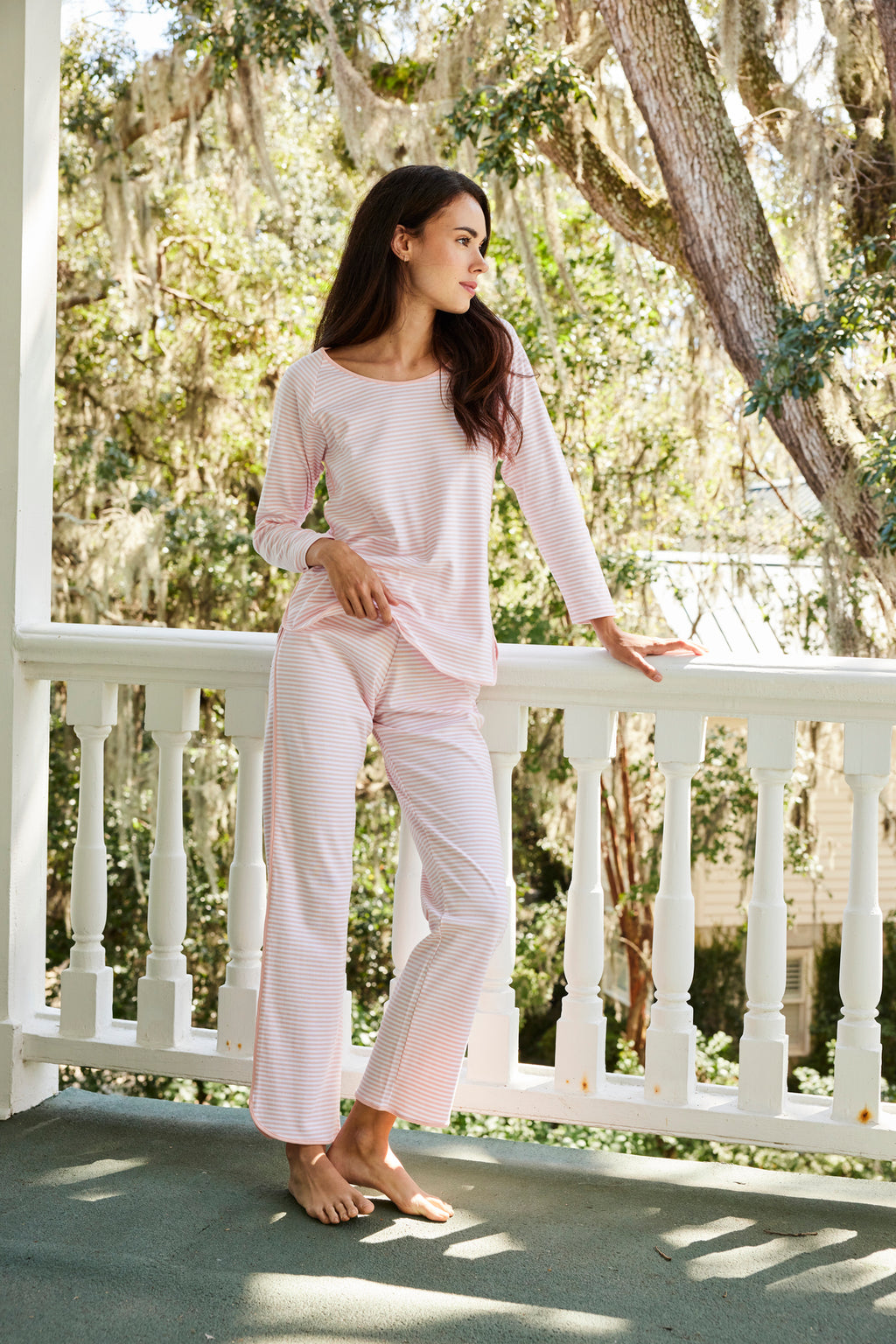 A woman in the Pima Long Sleeve Bundle in English Rose Stripe stands on a porch, one hand on the railing, gazing into the distance amid trees draped with moss and soft daylight.