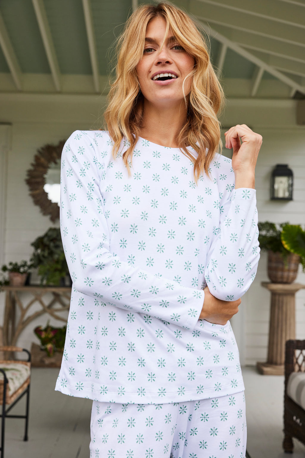 A woman smiles on a porch, wearing the Pima Crew Long-Long Set in Boxwood Moroccan Blockprint—a white crewneck top and matching high-waisted pants with a blue geometric pattern.