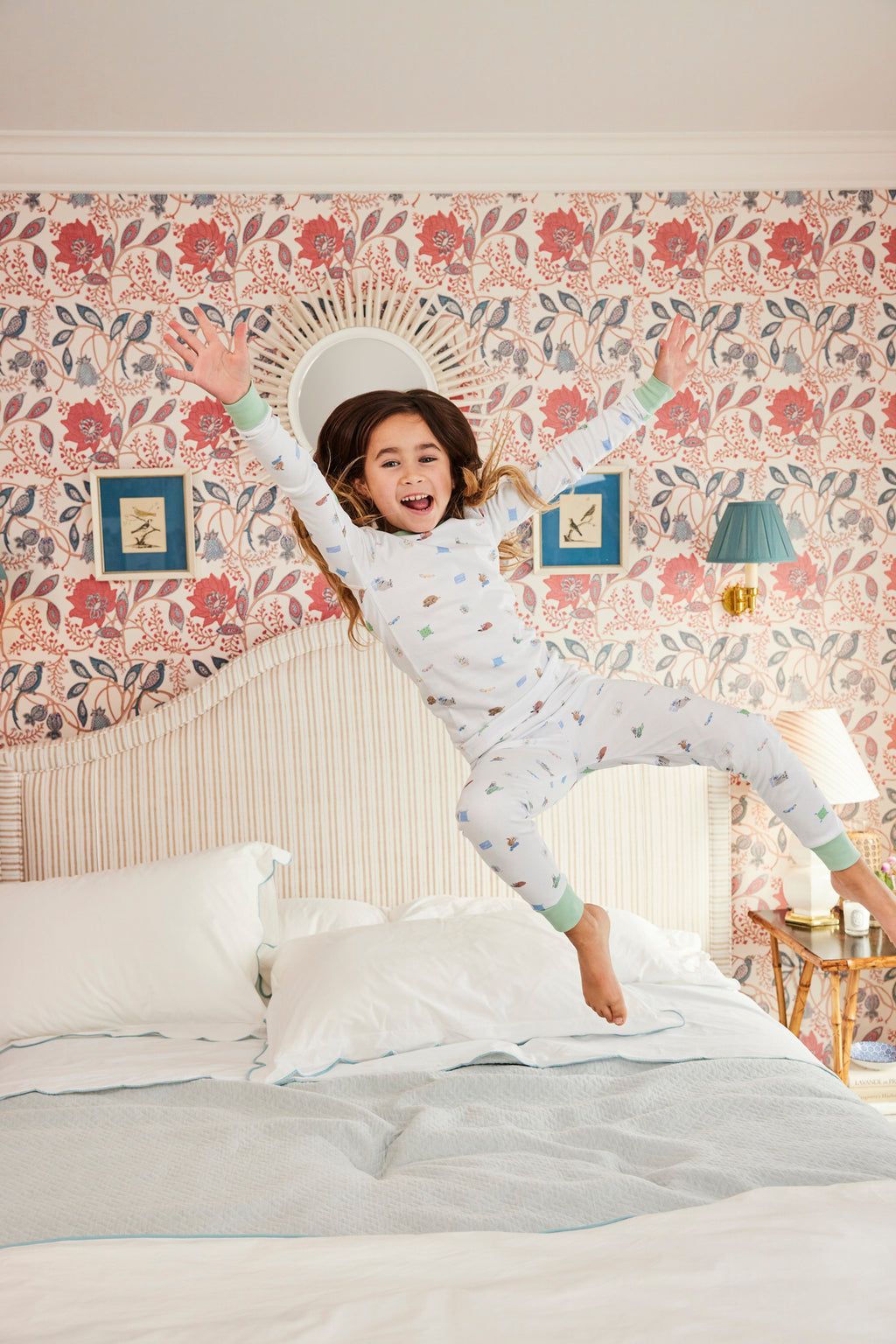 A young girl in the Kids Long-Long Set in Slumber Party smiles joyfully with arms raised as she jumps on a neatly made bed in a vibrant, floral-themed bedroom.
