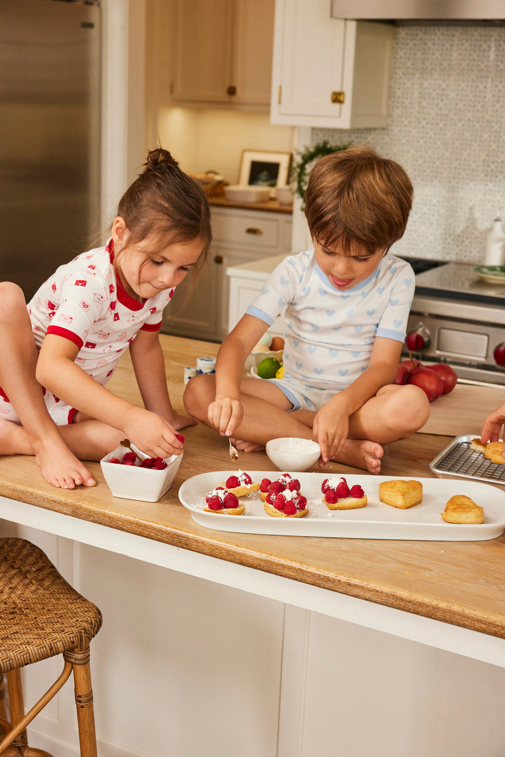 Two children wearing the Kids Shorts Set in French Blue Heart sit on a kitchen counter, smiling and focused as they make desserts with whipped cream and berries, surrounded by bowls and ingredients.