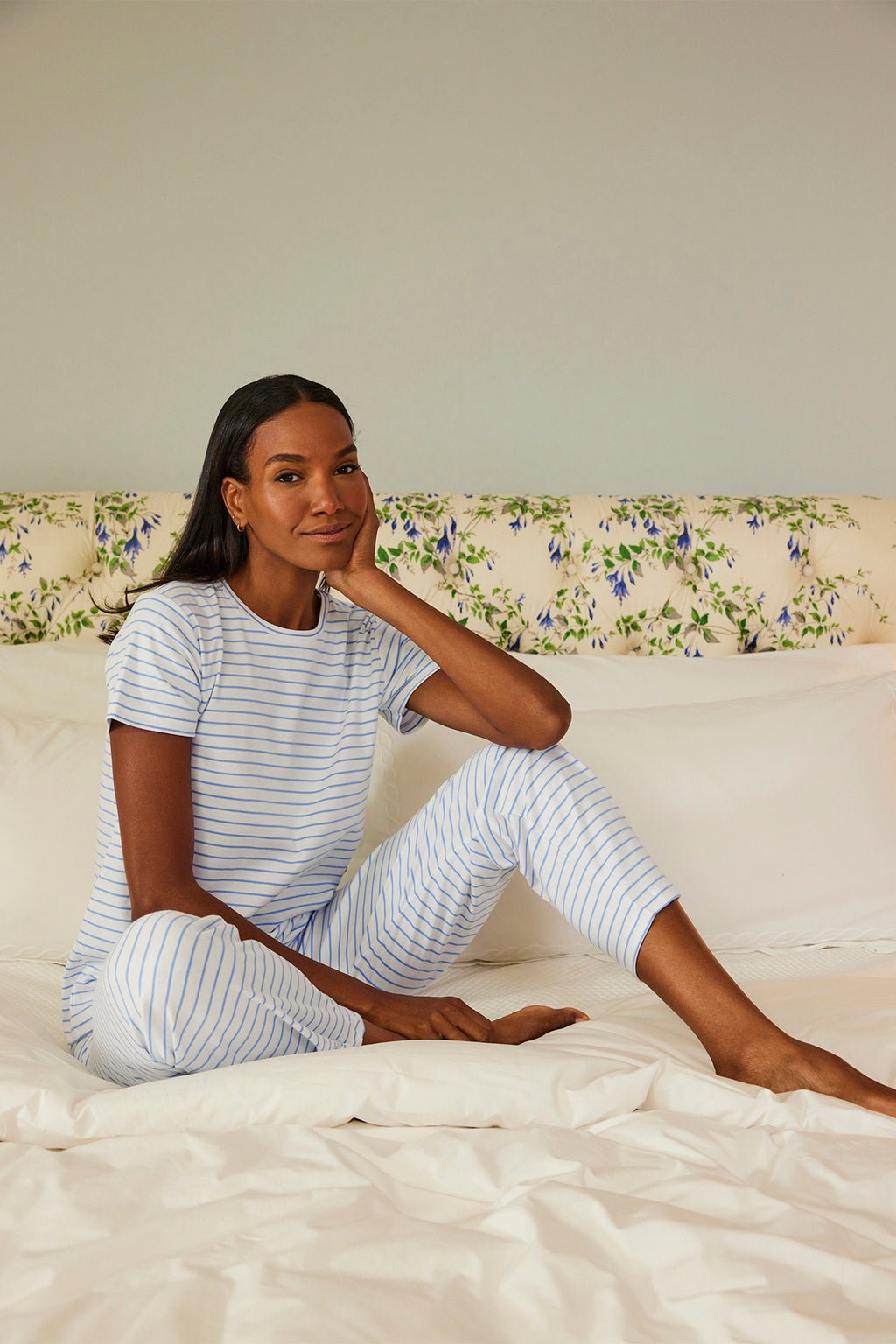 A woman wearing the Pima Crew Short-Long Set in Baltic Blue relaxes on a bed with white sheets, leaning on one hand. The bed’s floral-patterned headboard adds to the calm, cozy atmosphere.