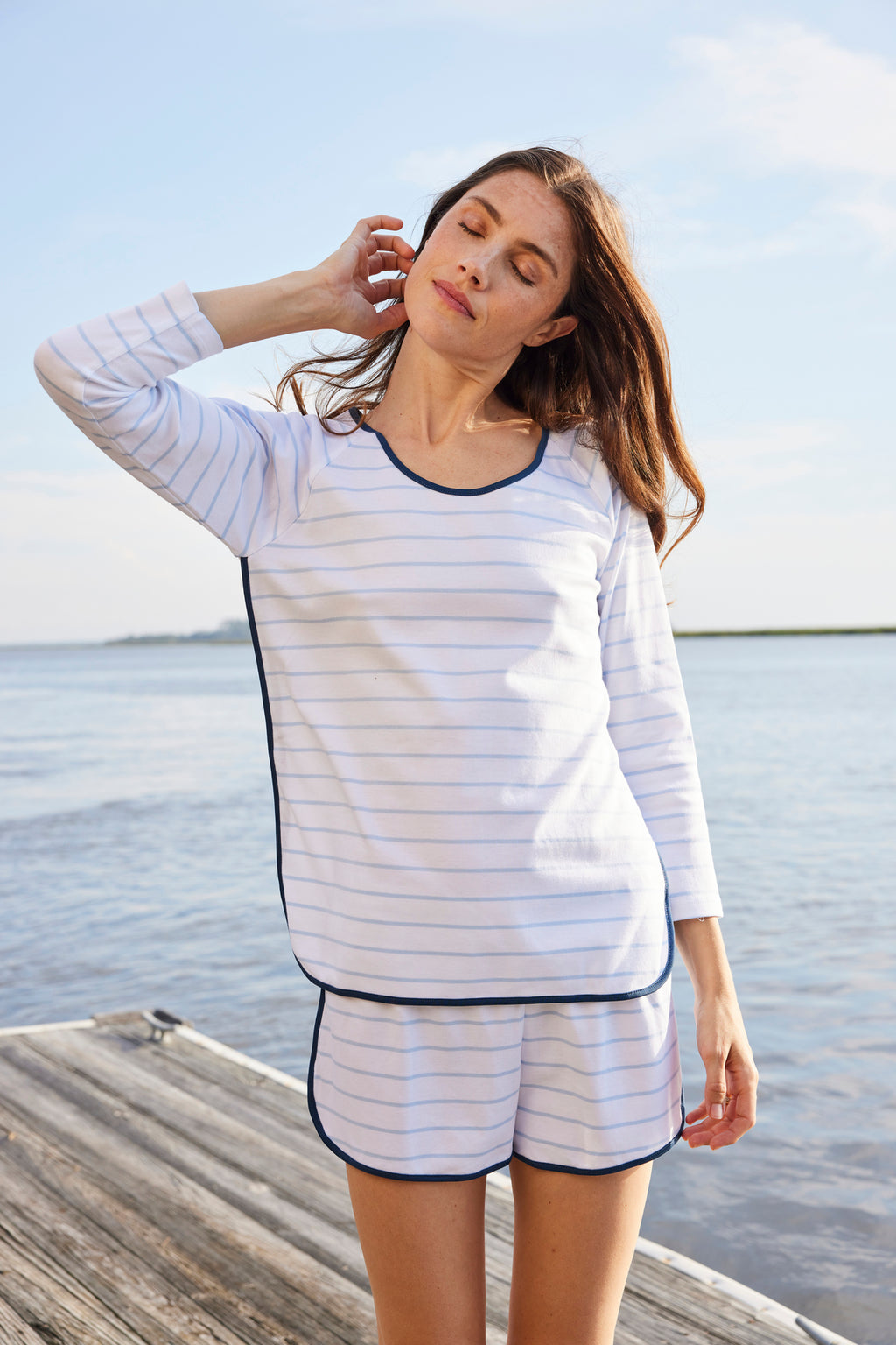 A woman stands on a wooden dock by the water, wearing the Pima Long-Short Set in Marine—100% Pima Cotton with thin blue Breton stripes. With one hand to her neck and eyes closed, she looks relaxed under a clear sky.