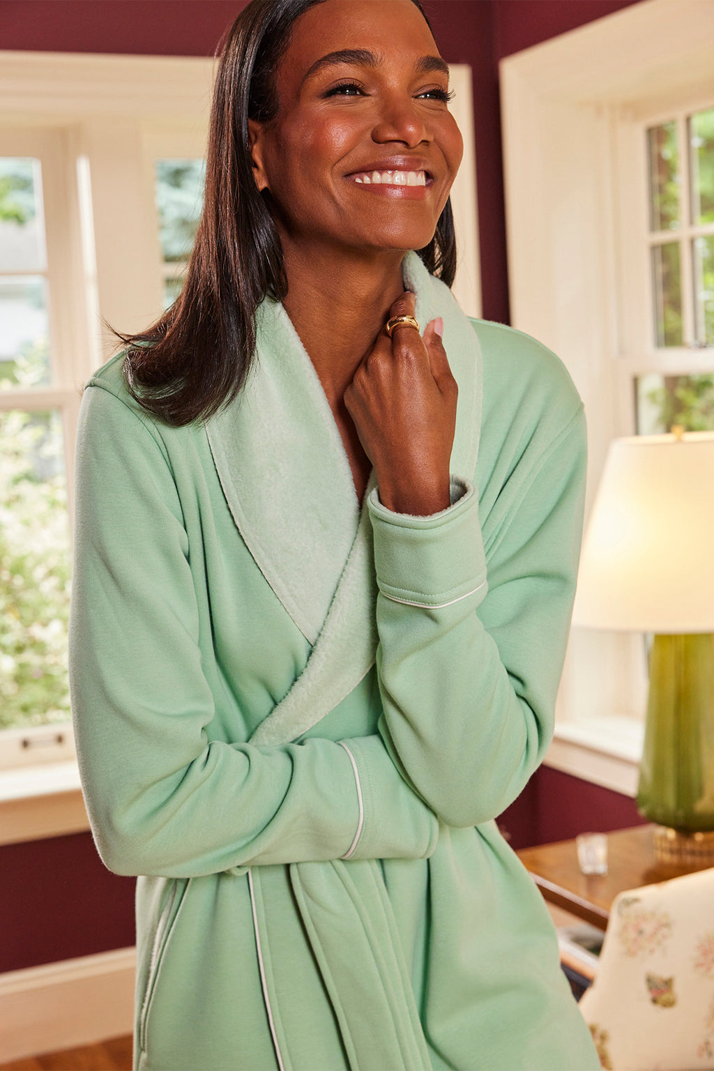 A woman smiles warmly indoors wearing the Cozy Robe in Parisian Green with white trim, posing with one hand near her collar as natural light streams through the windows behind her.