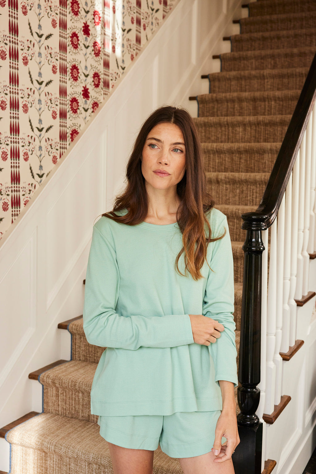 A woman in the Pointelle Shorts Bundle in Parisian Green Solid stands by a carpeted staircase with a floral wallpapered wall in the background.