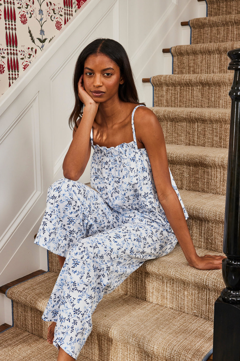A woman in a Pima Ruffle Pants Set in Serene Blue Botanical Vines sits on carpeted stairs, resting her chin on her hand and facing the camera, with patterned wallpaper and a black stair railing in the background.