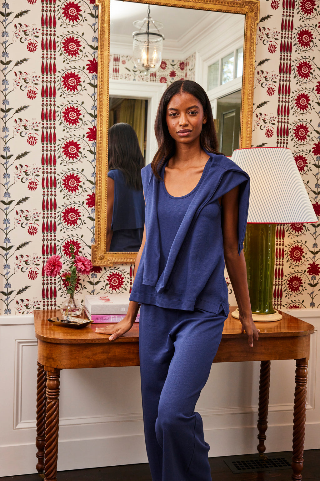 A woman wearing the Pointelle Pants Bundle in English Navy stands by a wooden table with flowers and a lamp, in a room featuring floral wallpaper and a large mirror.
