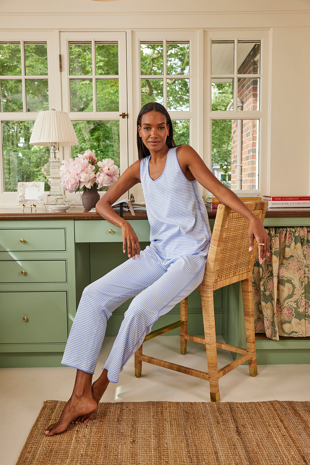 A woman in the Pima Tank-Long Set in Hydrangea, crafted from 100% Pima cotton, sits on a woven chair in a cozy room with green cabinets, a desk, flowers, books, and large windows revealing trees outside.