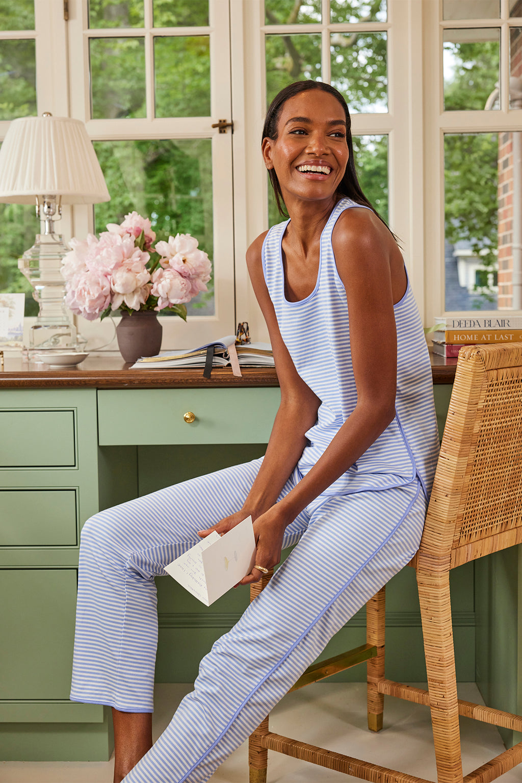 A woman in the Pima Tank-Long Set in Hydrangea, made of 100% Pima cotton in light blue and white stripes, sits on a wicker chair by a green desk with pink flowers and a lamp, smiling while holding a book near large windows.