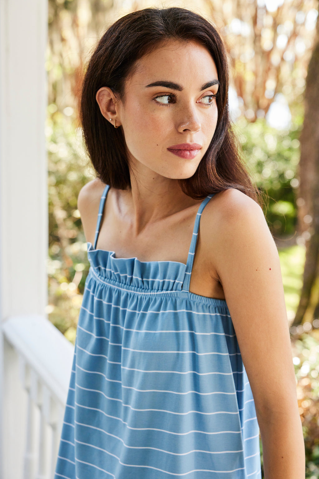 A woman in a Pima Ruffle Pants Set in Serene Blue Boating Stripe stands outdoors, gazing to the side amid vibrant greenery and gentle sunlight.