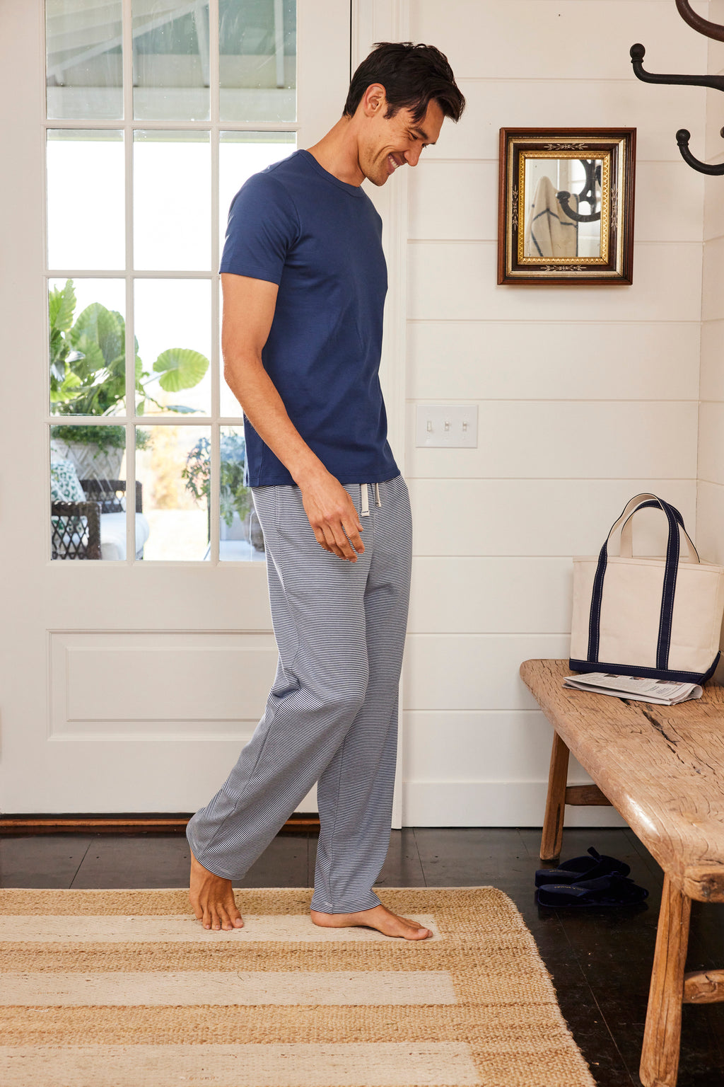 Barefoot on a woven rug in a sunlit, cozy room with a bench and striped bag, a man wears the Mens Pima Sleep Short-Long Set in True Navy with gray lounge pants. Perfect for relaxing comfort.