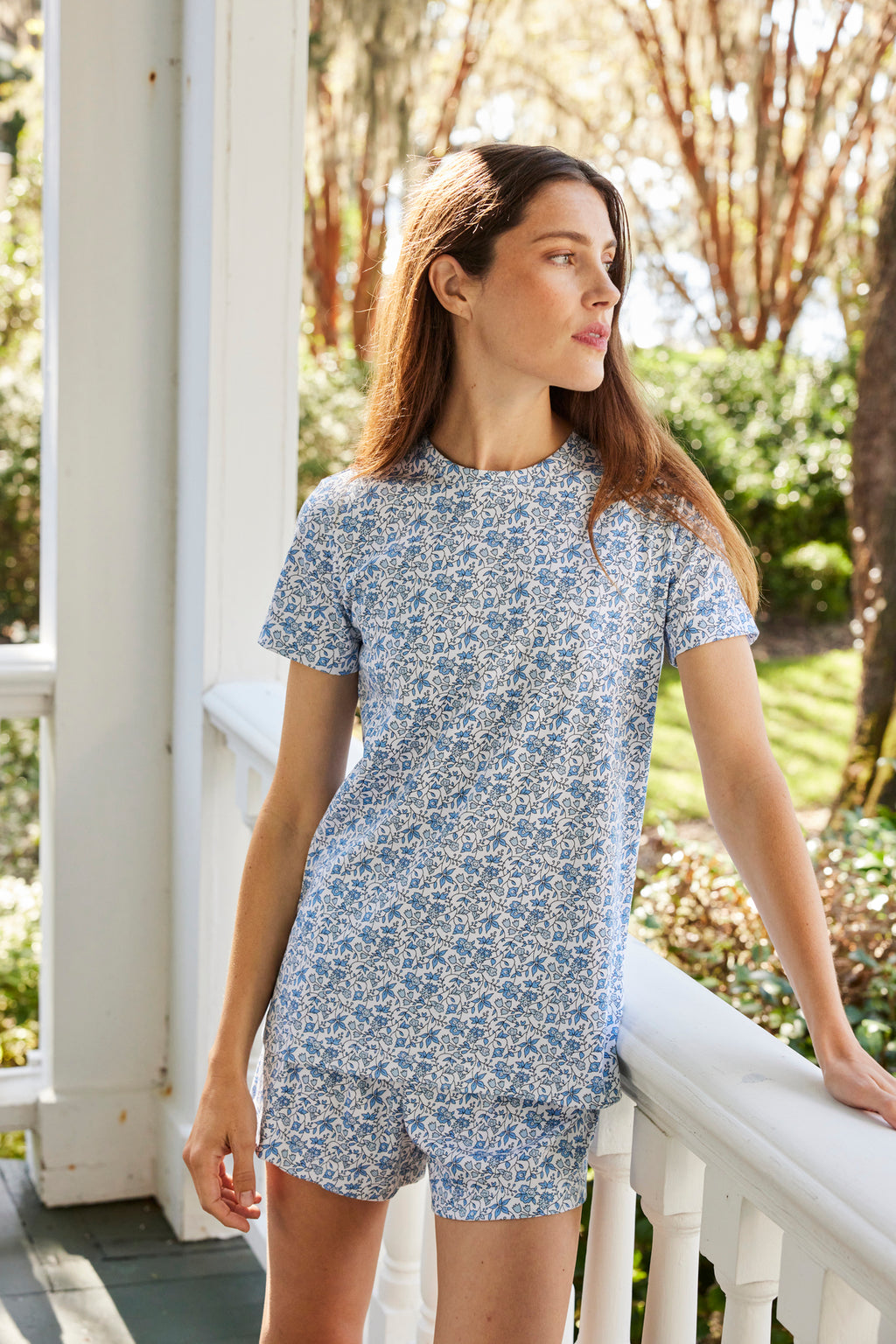 Wearing the Pima Crew Shorts Set in Delft Blue Garden Floral, a woman stands on a porch, resting one hand on the railing and looking to the side with greenery and sunlight in the background.