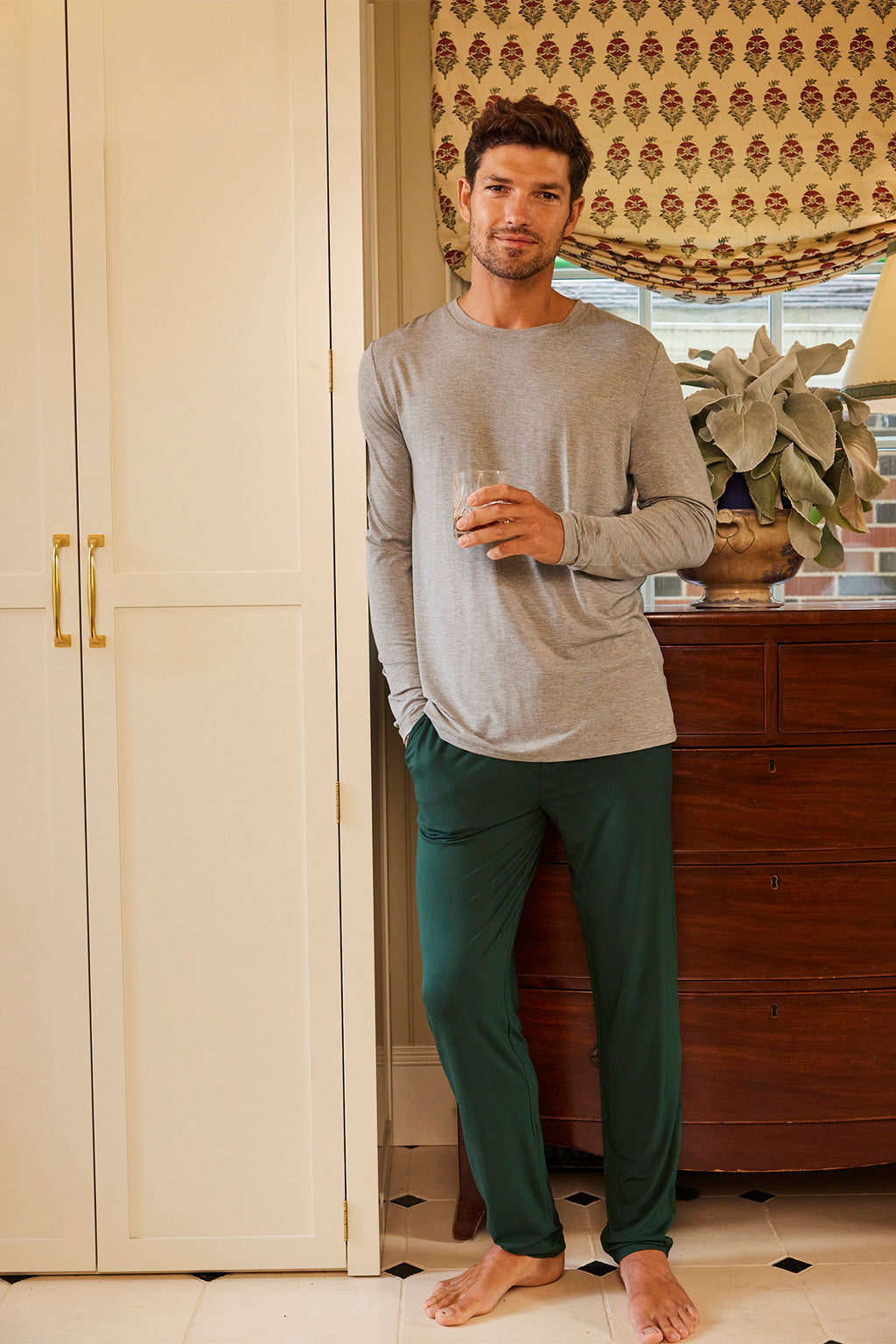 A man stands indoors, leaning against a cream-colored cabinet. He wears a gray long-sleeve shirt and Mens Bamboo Lounge Pants in Conifer with an elastic waistband, holding a glass with a plant and chest of drawers in the background.