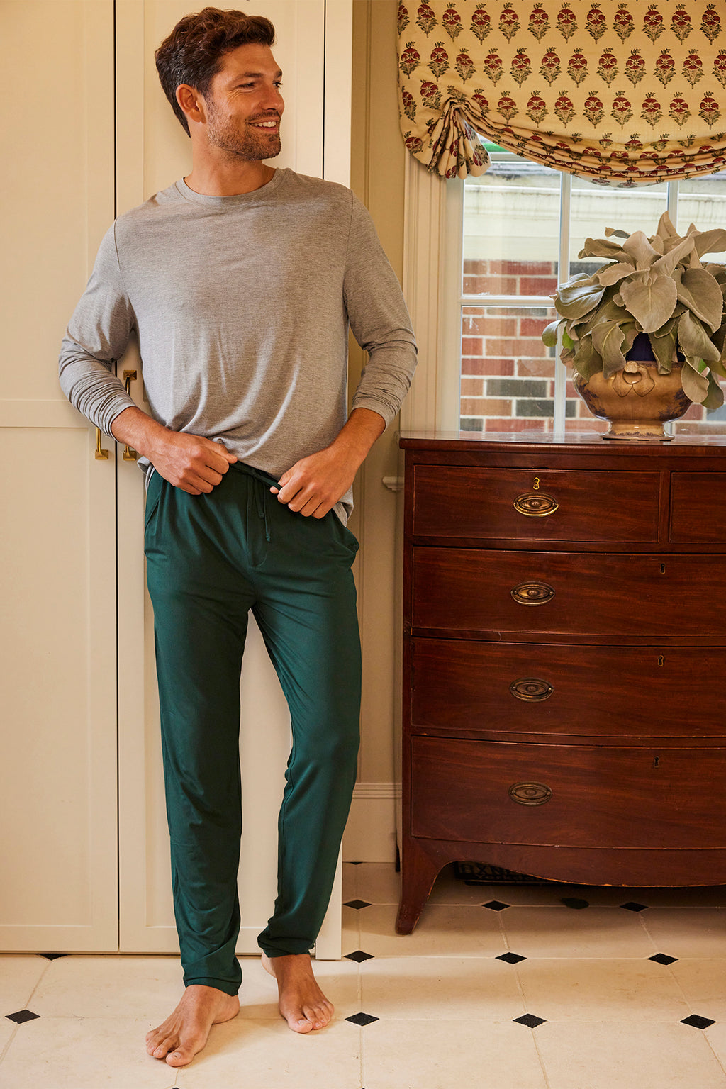 A man stands indoors, smiling and wearing a light gray long-sleeve shirt and Mens Bamboo Lounge Pants in Conifer with an elastic waistband, leaning casually against a kitchen counter beside a wooden dresser and a window with a patterned shade.