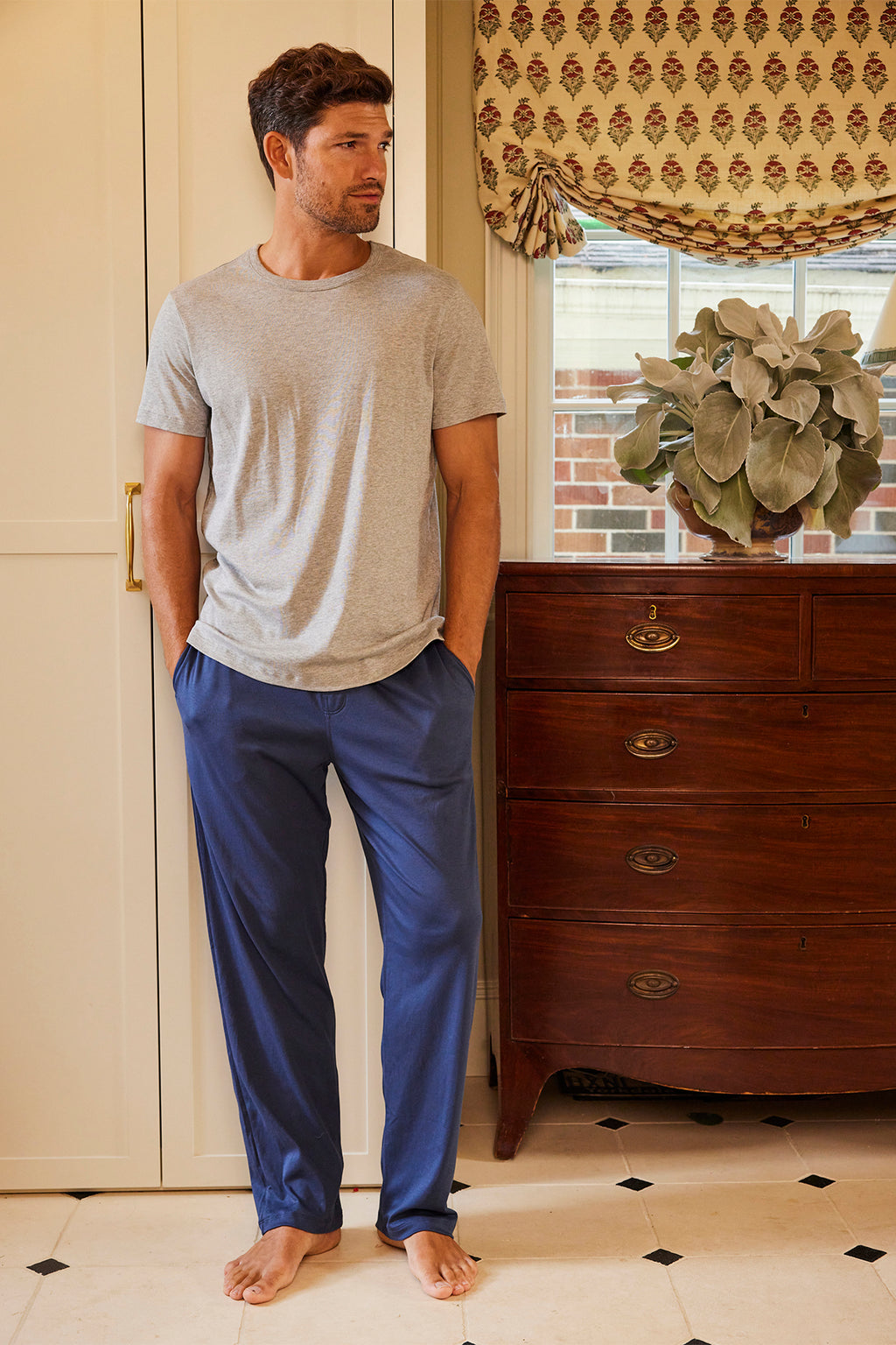 A man stands indoors on a tiled floor, wearing the Mens Pima Sleep Bundle in Graphite Helm Stripe. Hands in pockets, he looks to the side; behind him are a wooden dresser, a plant, and a window with a patterned curtain.