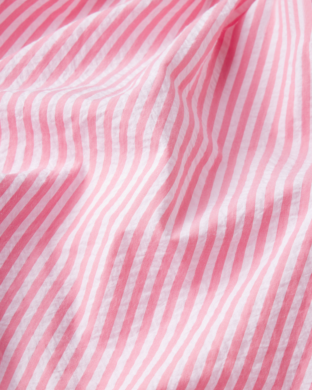 A close-up of pink and white vertically striped Sunkissed Coral Seersucker fabric with soft folds, featured in the Alice Shorts Set in Sunkissed Coral Seersucker, highlights its textured look—perfect for this stylish set.