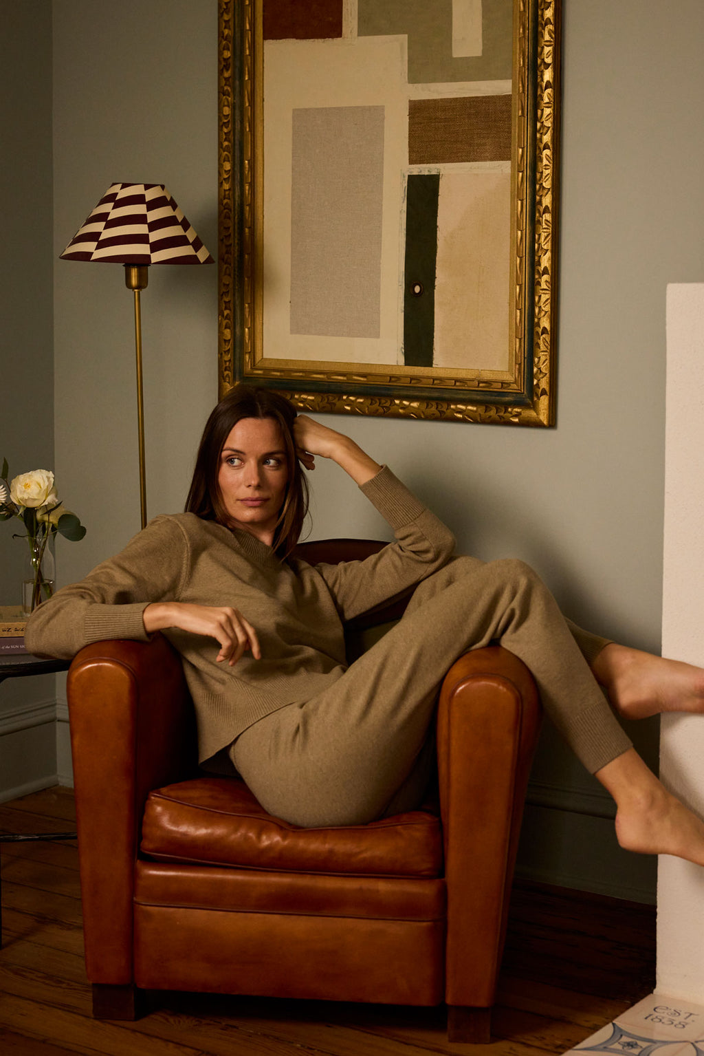 A woman in the Vesper Crew Neck Set in Sandstone Heather relaxes in a brown leather armchair, one arm on the chair and the other on her head. Behind her are a framed abstract painting and a lamp with a striped shade.