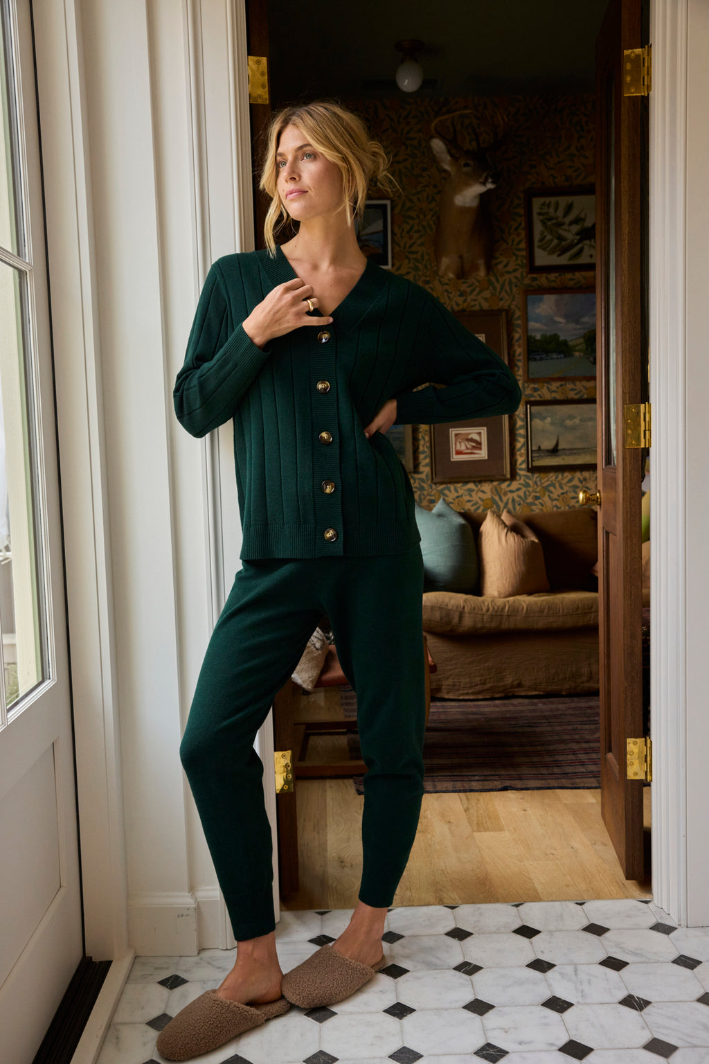 A woman stands in a doorway wearing the Vesper Jogger Set in Conifer and tan slippers. Behind her, a cozy room features a sofa, patterned wallpaper, and framed artwork on the walls.