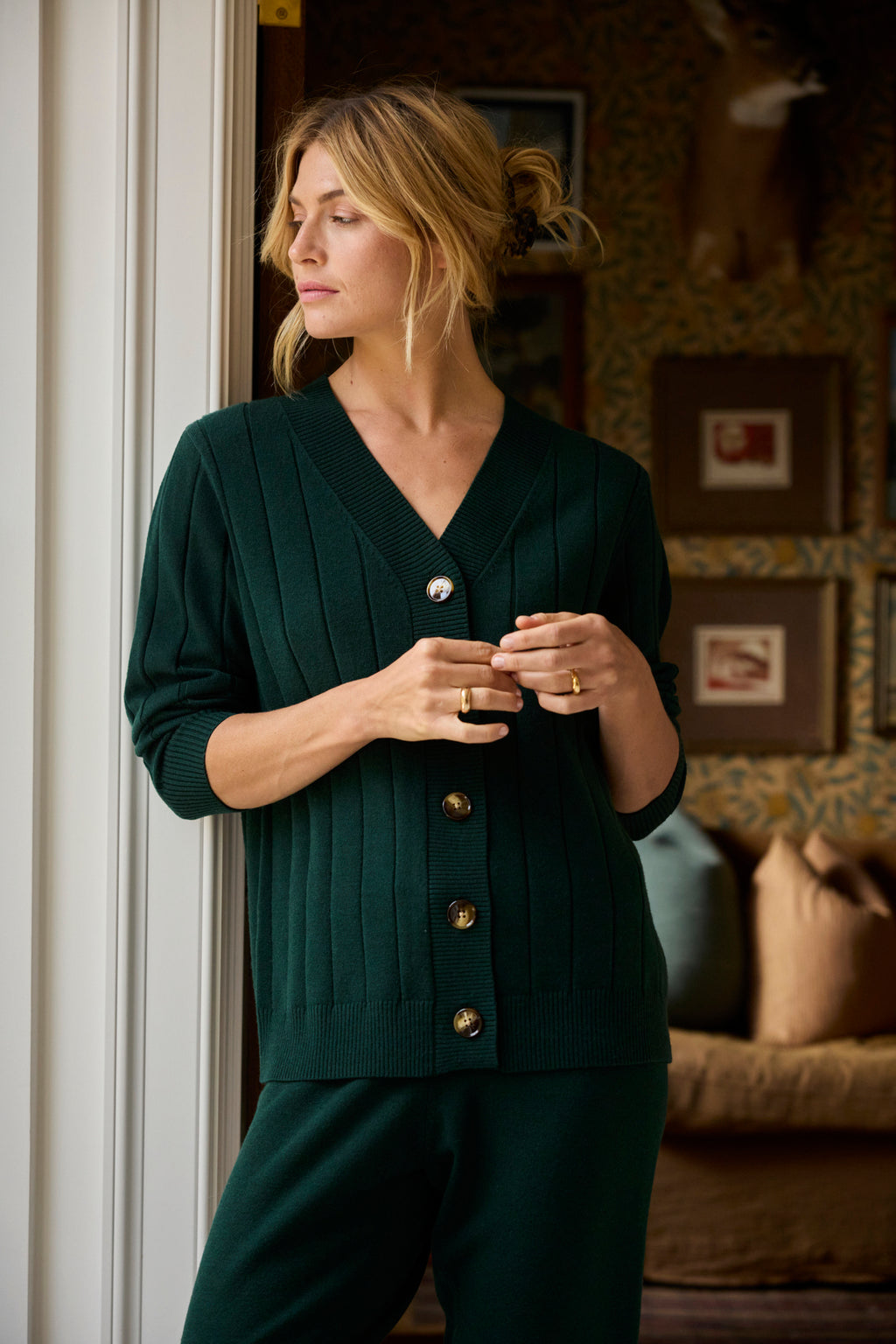 A woman wearing the Vesper Jogger Set in Conifer stands by a window, gazing to the side in a cozy, warmly-lit room with framed art on the wall.