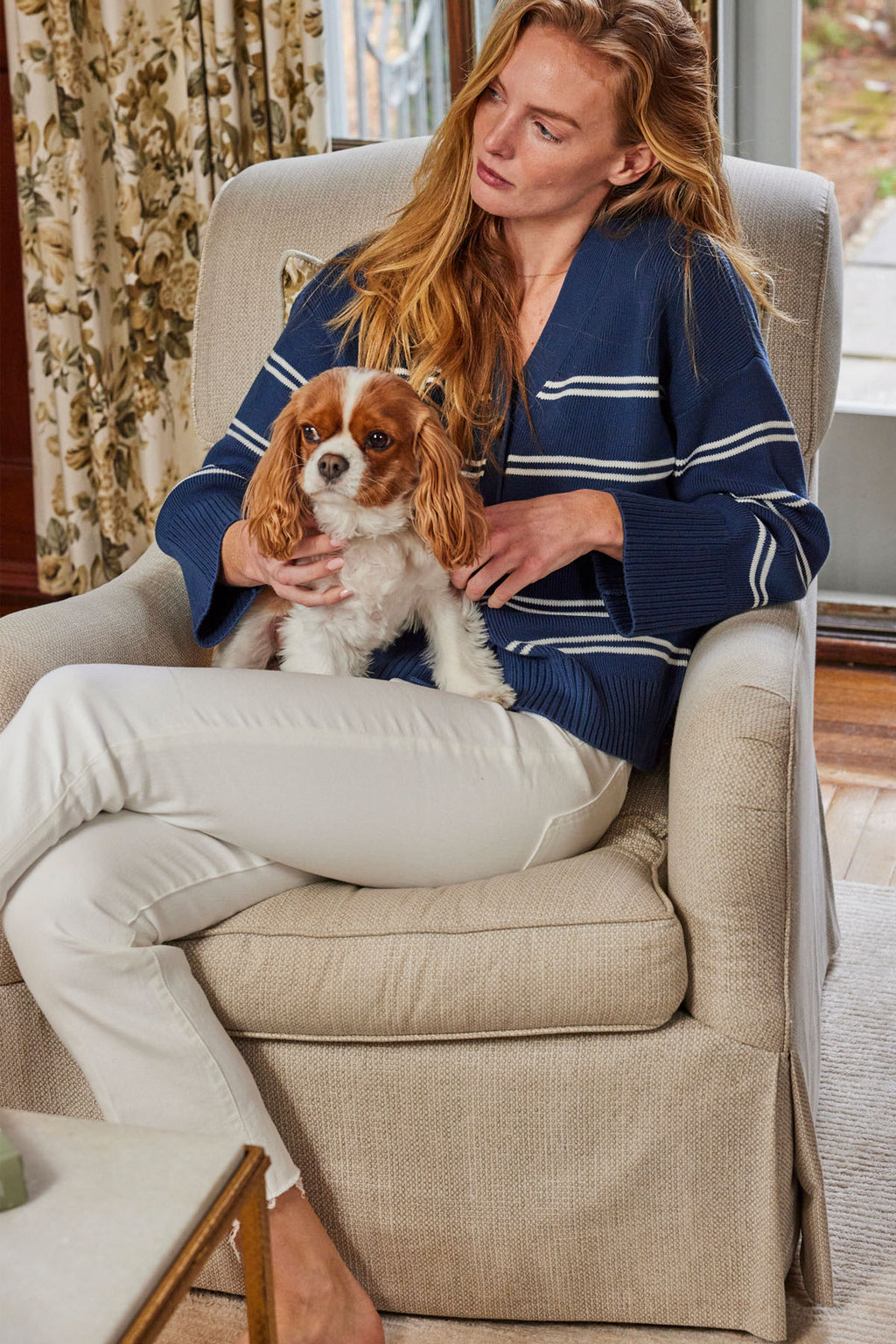 A woman sits indoors in a beige armchair, wearing the Nora Cotton Cardigan in Navy Ivory Stripe and white pants, gently holding a small brown and white dog on her lap.