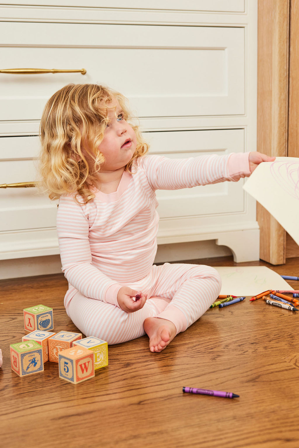 A young child wearing the Kids Long-Long Set in English Rose Stripe with an elastic waistband sits on a wooden floor, holding paper, with crayons and wooden alphabet blocks nearby, looking up and reaching out.