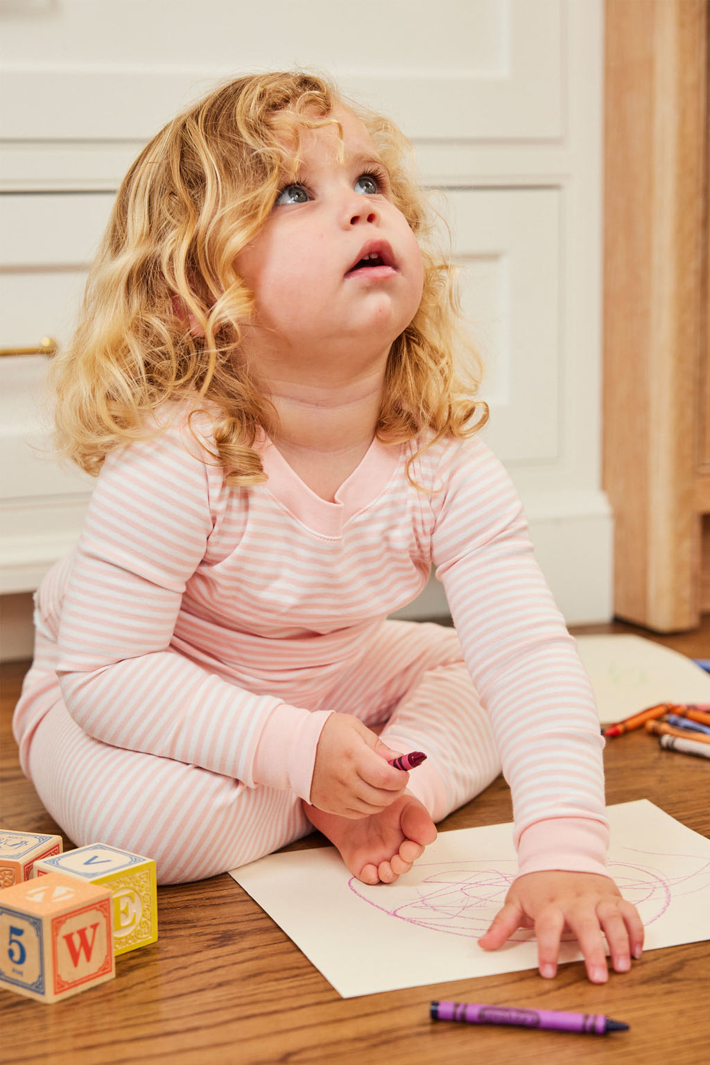 A young child in Kids Long-Long Set in English Rose Stripe pajamas made from soft Pima cotton sits on a wooden floor drawing with crayons, with colorful blocks and crayons scattered around.