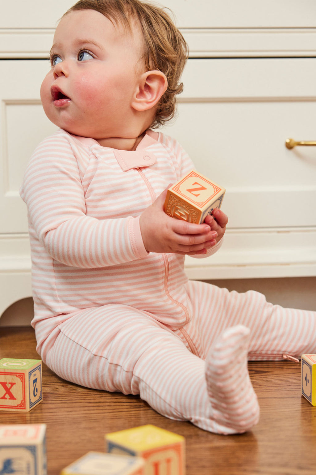 A baby in pink and white English Rose Stripe Baby Sleeper sits on a wooden floor, holding a wooden alphabet block and gazing upward, with other colorful blocks scattered nearby to showcase this cozy outfit.