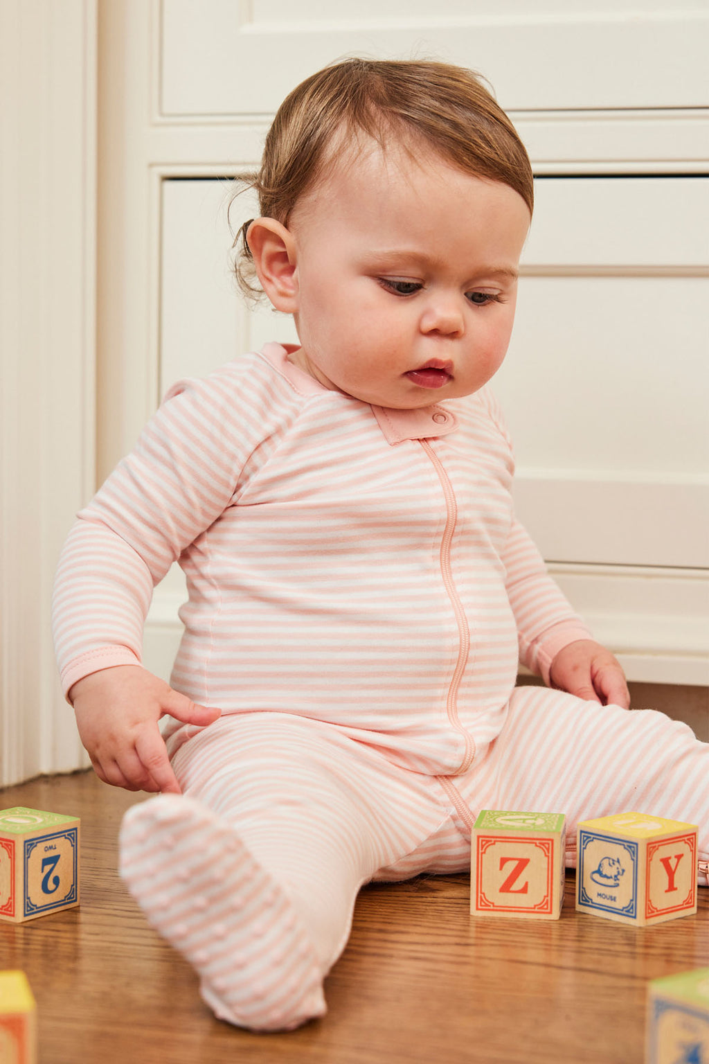 A baby wearing the Baby Sleeper in English Rose Stripe sits on a wooden floor, gazing down at colorful alphabet blocks.