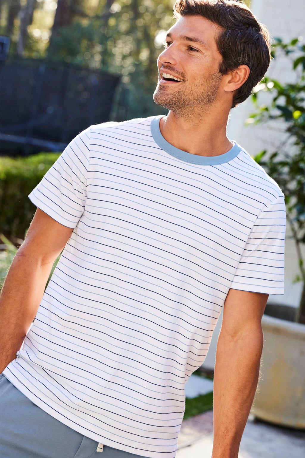 A man outdoors smiles, wearing the Mens Pima Sleep Shorts Set in Marlin Blue Nautical Stripe, featuring soft 100% Pima cotton and a light blue collar, with a blurred green and sunlit background.