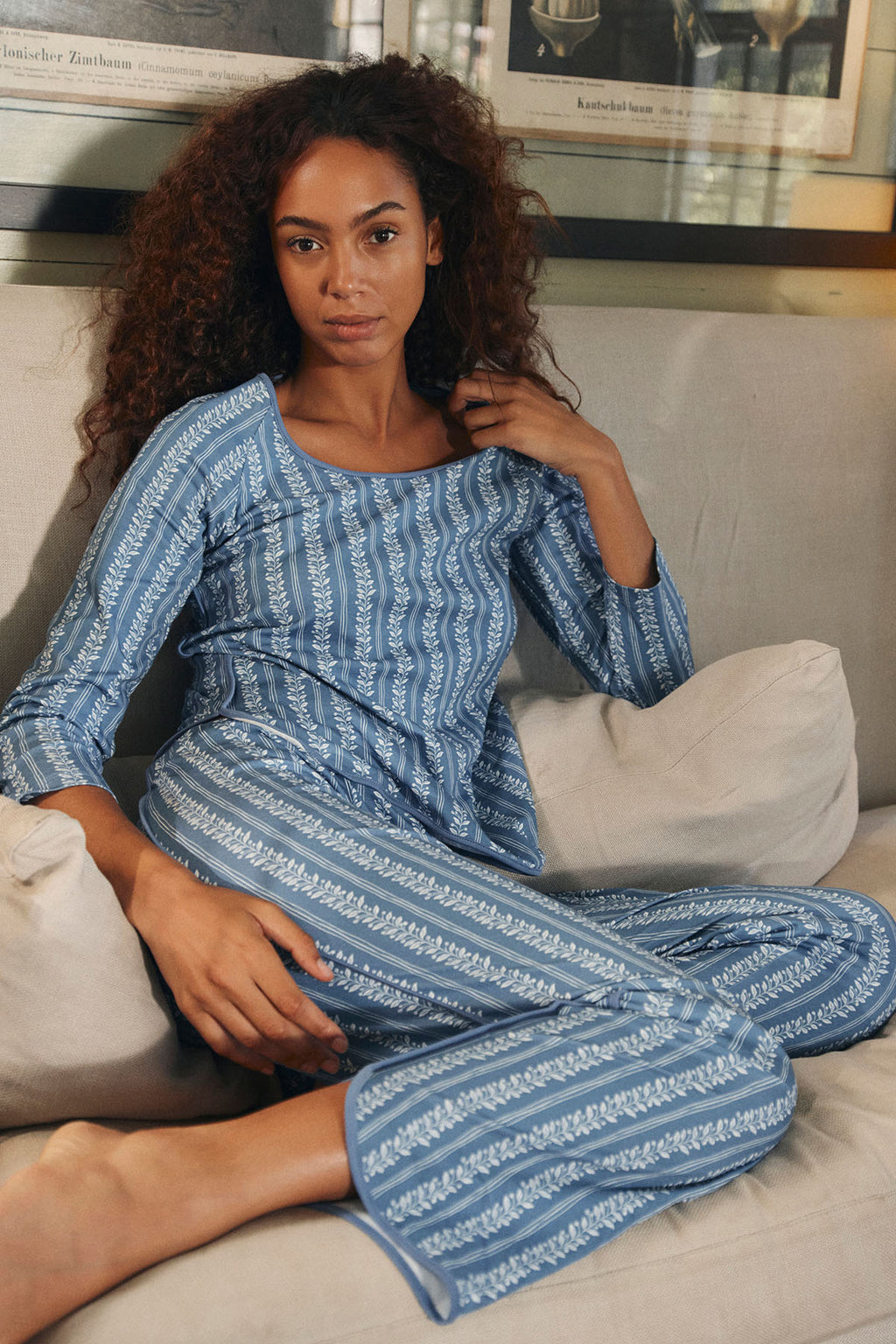 A woman wearing the Pima Long-Long Set in Seaport Jasmine Vine lounges on a cream-colored couch, looking relaxed in her matching blue long-sleeve top and pants with a white, leafy pattern. One arm rests on a cushion, the other gently raised.