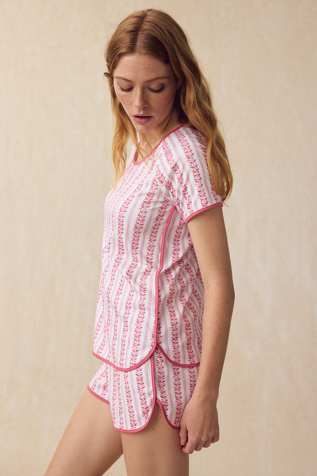 A woman looks down while wearing the Pima Shorts Set in Winterberry Jasmine Vine, a women’s sleepwear pajama shorts set with a white base, pink floral vine patterns, and pink trim, standing against a neutral background.