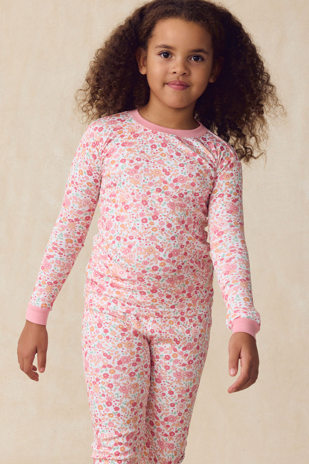 A young child wears the Kids Long-Long Set in Rosewater Vivian Floral, featuring a floral print with pink cuffs, standing against a light beige background. These pajamas offer both comfort and style for kids.