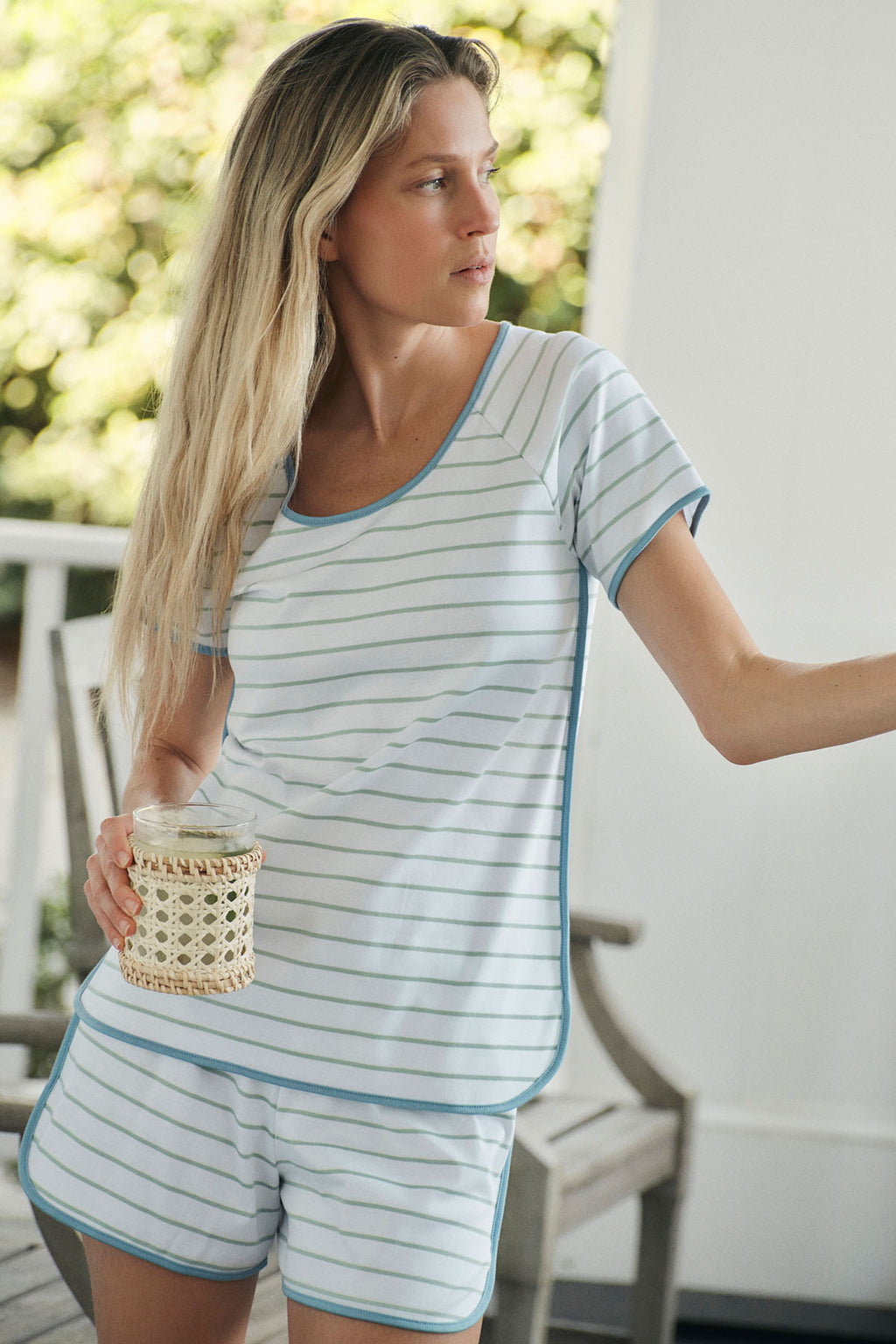 A woman in the Pima Shorts Set in Parisian Green Club Stripe stands on a porch, holding a woven cup and looking to the side. A wooden chair and lush greenery outside highlight the set’s breathable sleepwear comfort.