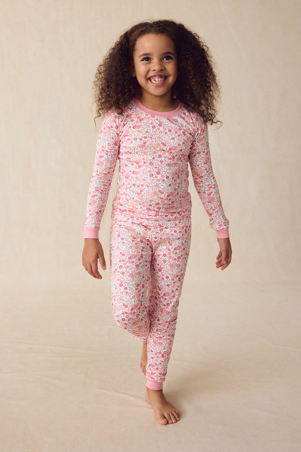 A young girl smiles in the Kids Long-Long Set in Rosewater Vivian Floral, featuring matching floral long-sleeve Pima cotton pajamas with pink cuffs, standing against a plain beige background.