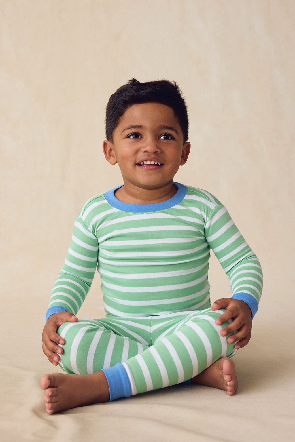 A young child sits on the floor, smiling in the Kids Long-Long Set in Field Green Hull Stripe. The pajama set features blue cuffs and a green and white stripe pattern, set against a soft, light beige background.