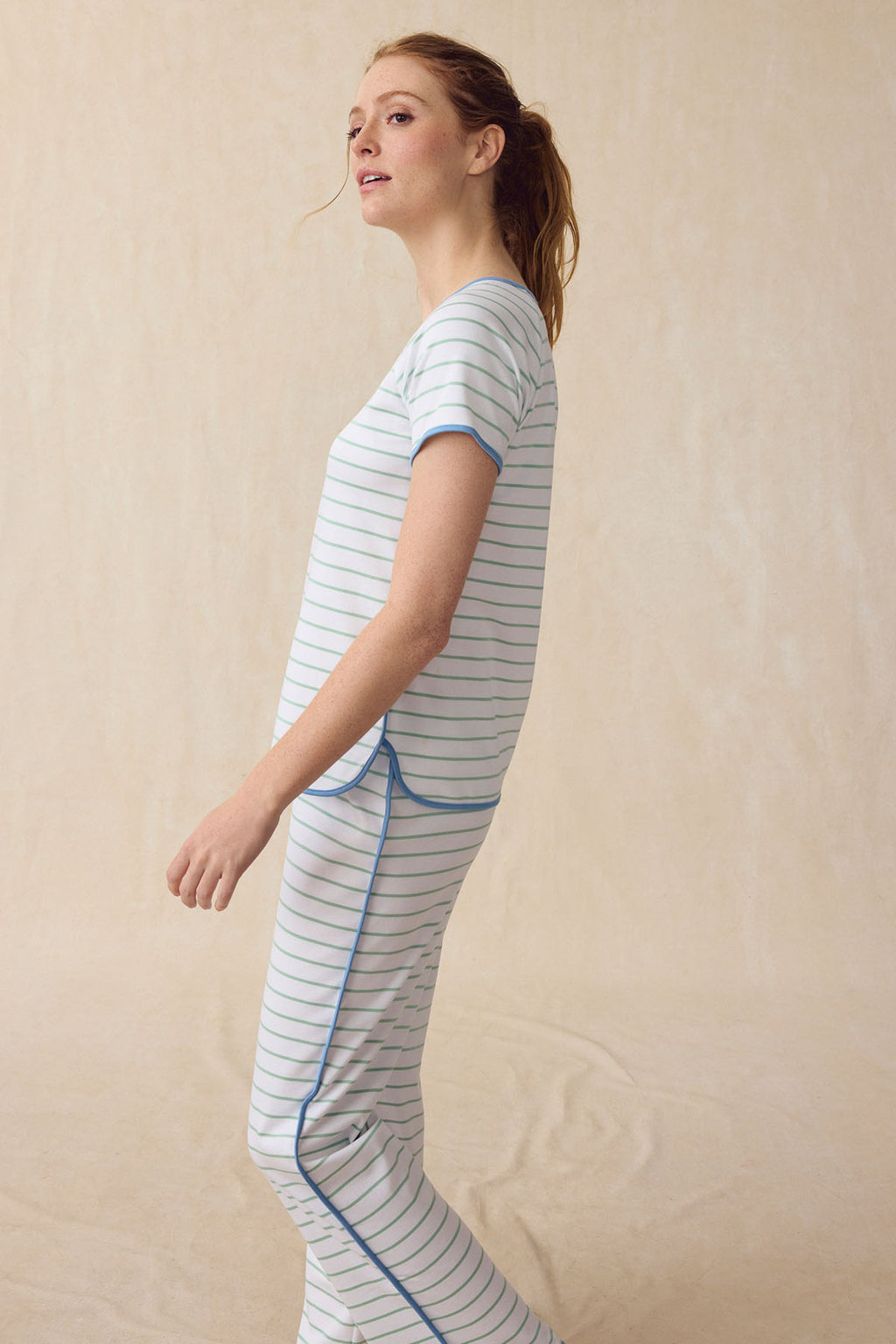 A woman with light skin and a ponytail models the Pima Short-Long Set in Parisian Green Club Stripe, featuring a breathable short-sleeve top and matching shorts. She stands sideways against a beige backdrop, gazing slightly upward.
