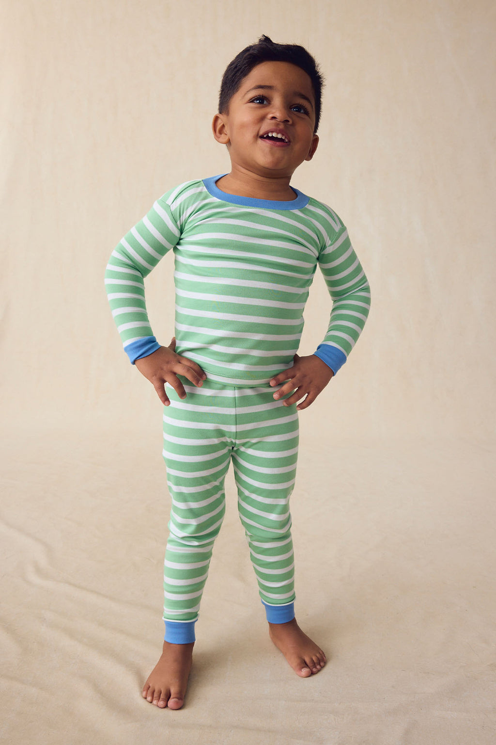 A young child smiles in the Kids Long-Long Set in Field Green Hull Stripe, featuring green striped Pima cotton pajamas with blue cuffs, standing against a neutral beige background.