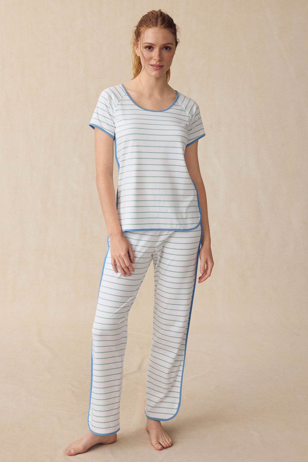 A woman stands on a neutral background, wearing the Pima Short-Long Set in Parisian Green Club Stripe—a breathable pajama set with thin green stripes, blue trim, a short-sleeve top, and long pants.