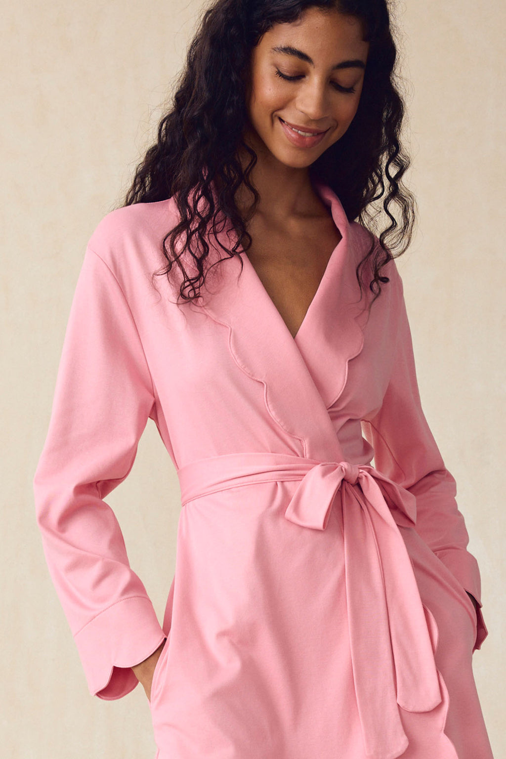 A woman smiles in the Pima Scallop Robe in Rosewater, featuring a scalloped lapel and belted waist, against a neutral background.