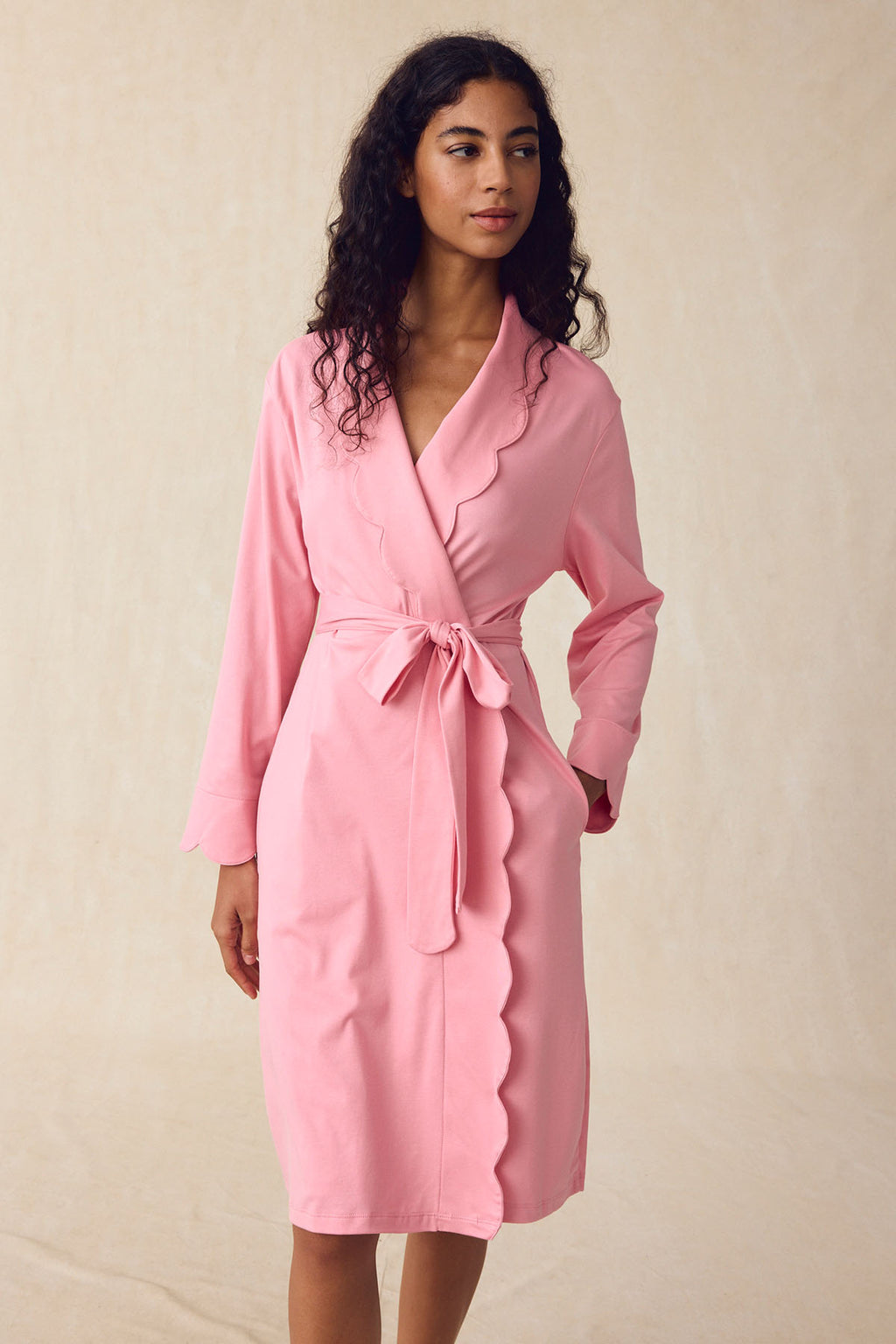 A woman wears the Pima Scallop Robe in Rosewater, a light pink lounge robe with scalloped edges, a scalloped lapel, and a tie belt, standing against a plain beige background.