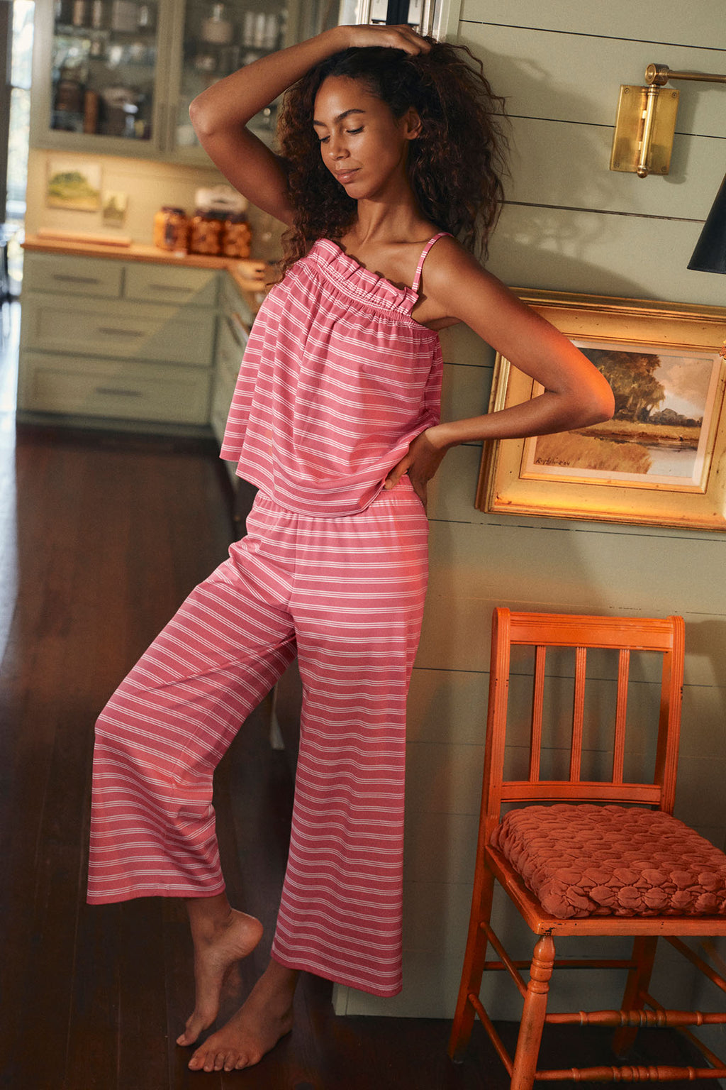 A woman stands indoors with one hand on her hip and eyes closed, wearing the Pima Ruffle Pants Set in Winterberry Triple Stripe. She leans against a wall near a wooden chair in a cozy, warmly lit room.