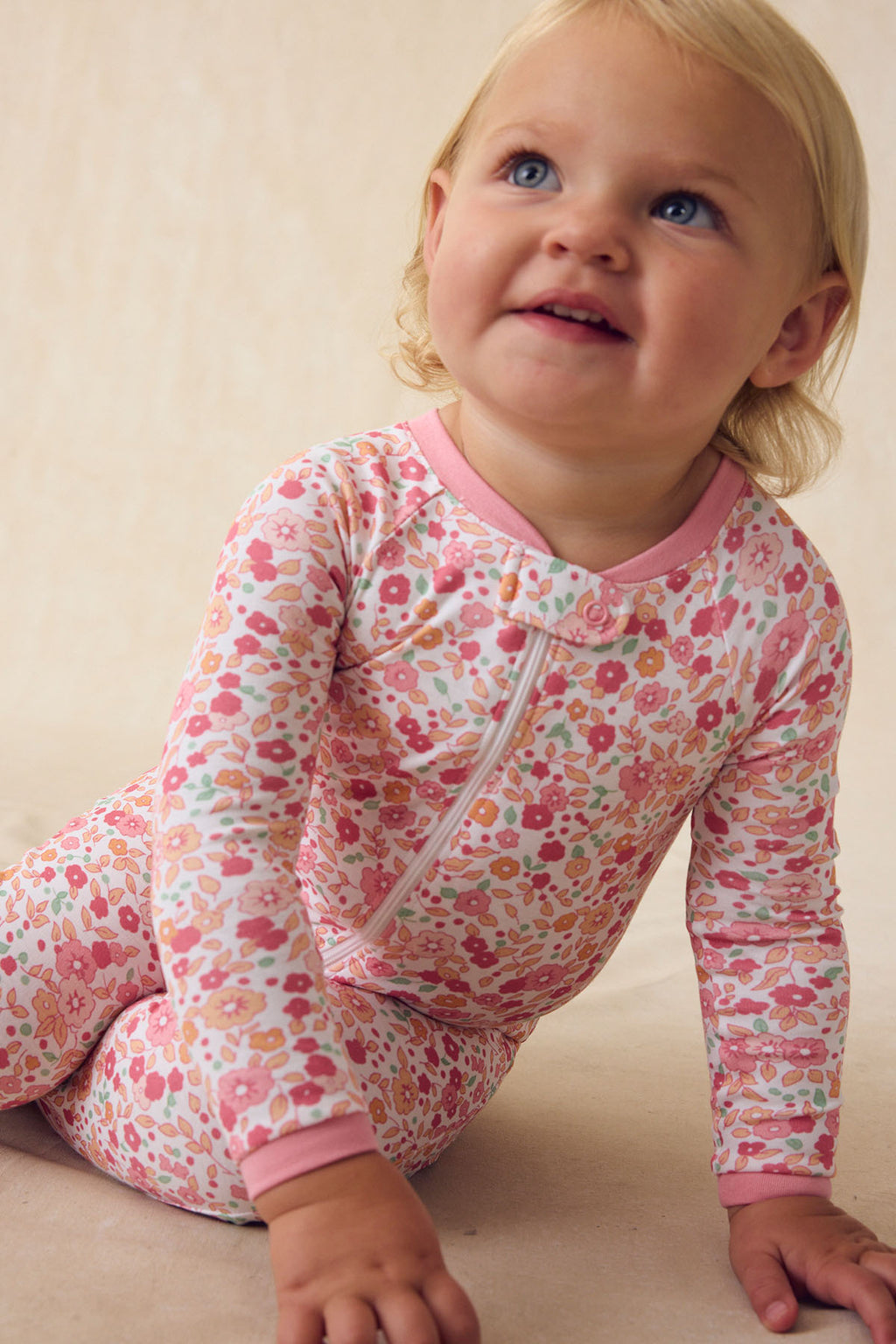 A blue-eyed toddler sits on the floor, smiling and looking up, wearing the Baby Sleeper in Rosewater Vivian Floral made from soft Pima cotton with pink trim and a dual zipper.