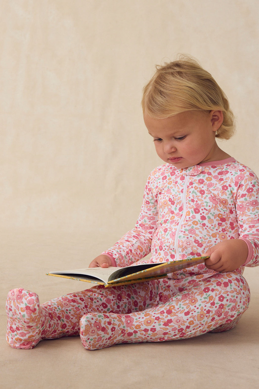 A young child wearing the Baby Sleeper in Rosewater Vivian Floral sits on a soft surface, intently looking at an open hardcover book in their lap.