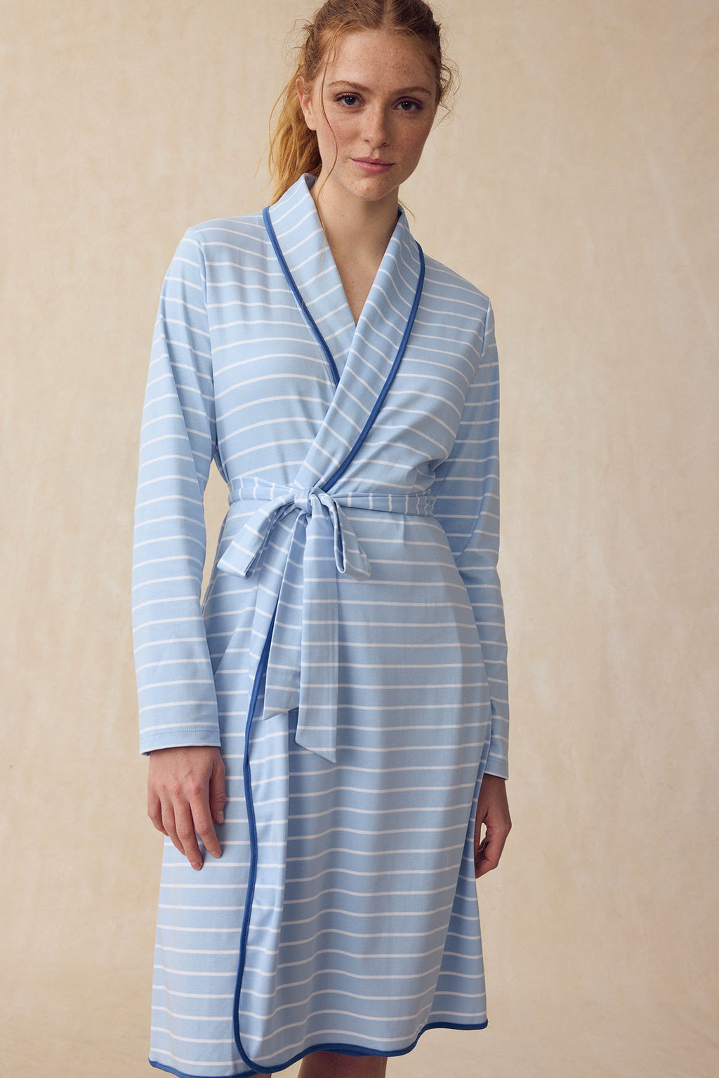 A woman stands against a beige background wearing the Pima Robe in French Blue Club Stripe, featuring long sleeves, white horizontal stripes, blue trim, and a matching belt.