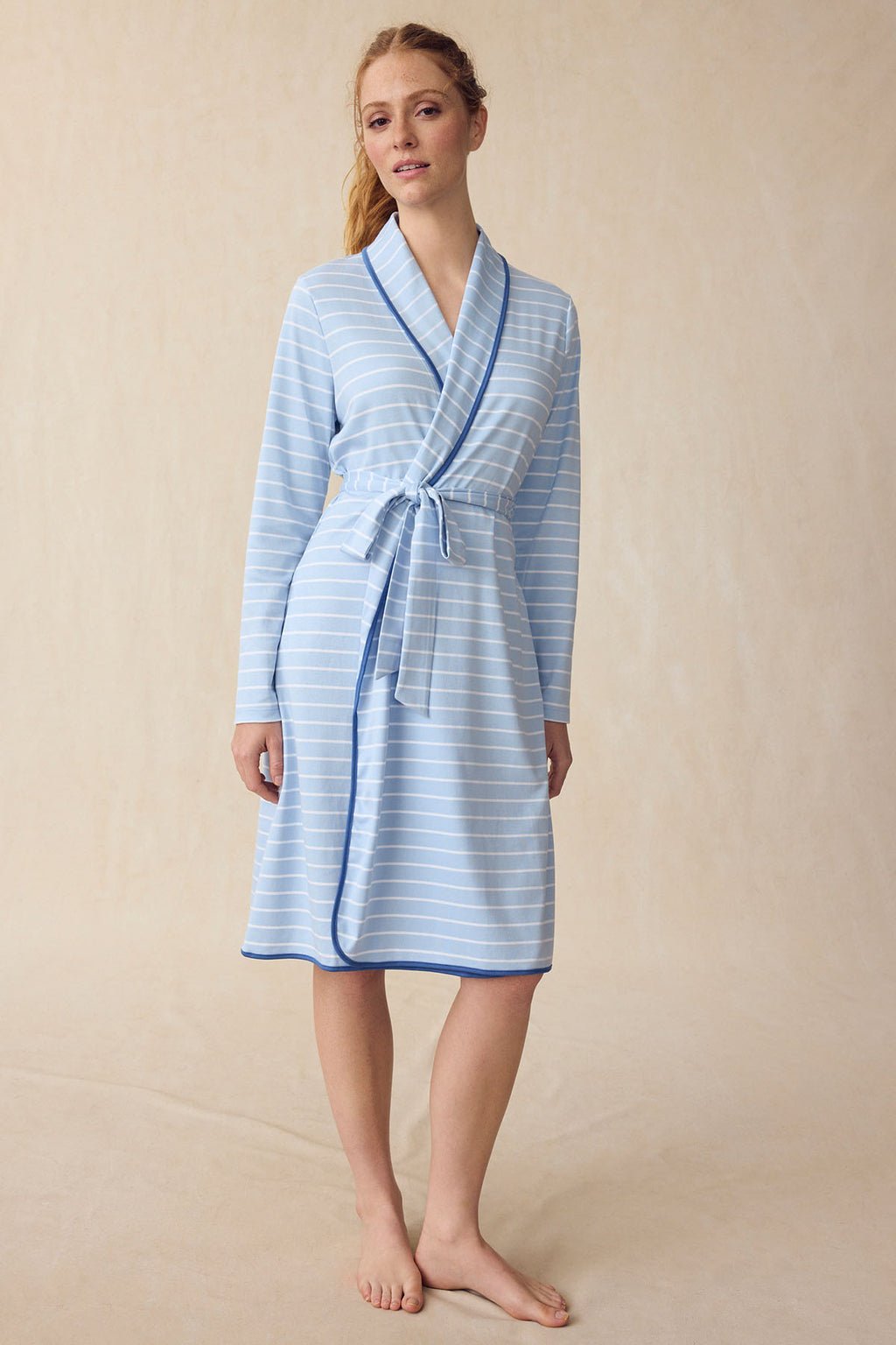 A woman wears the Pima Robe in French Blue Club Stripe—light blue with thin white stripes and a matching belt—knee-length, against a neutral beige background.