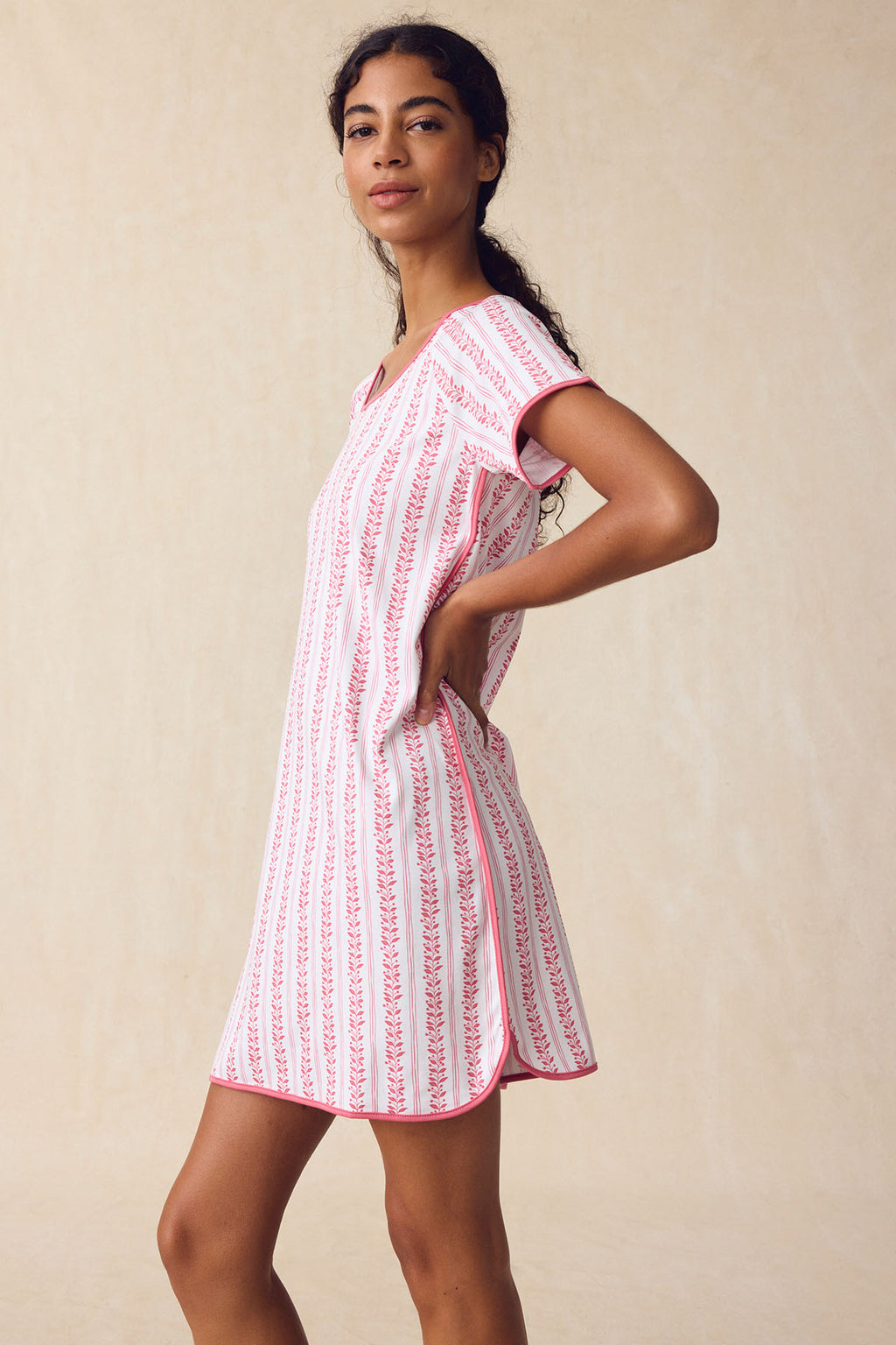 A woman stands sideways, looking at the camera, wearing the Pima Nightgown in Winterberry Jasmine Vine—a short-sleeved dress with vertical pink patterns—against a neutral, plain background.