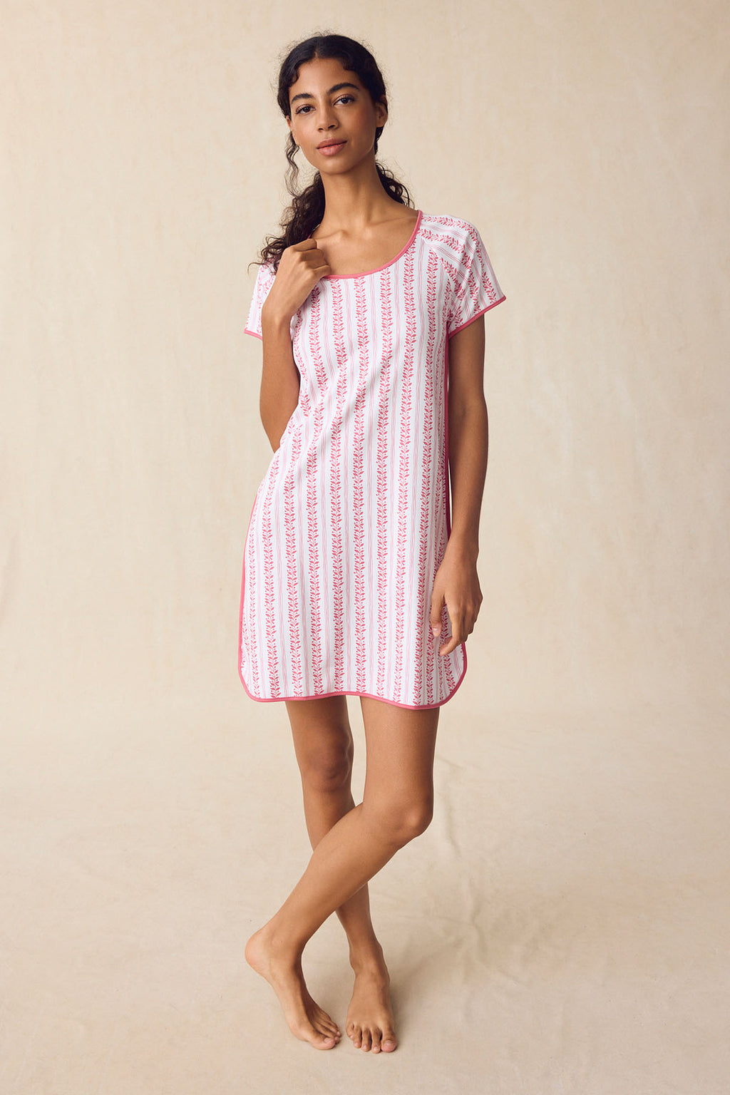 A woman stands on a neutral background wearing the Pima Nightgown in Winterberry Jasmine Vine—a short-sleeved, knee-length piece featuring pink vertical patterns and pink trim. She has a relaxed expression.