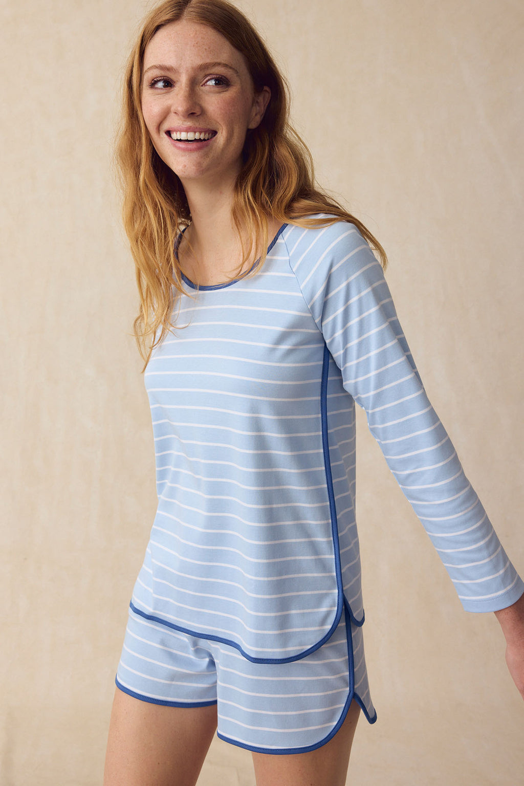 A woman smiles in the Pima Long Sleeve Bundle in French Blue Club Stripe—featuring a light blue and white long-sleeve top and matching elastic-waist shorts—while standing against a neutral beige background.