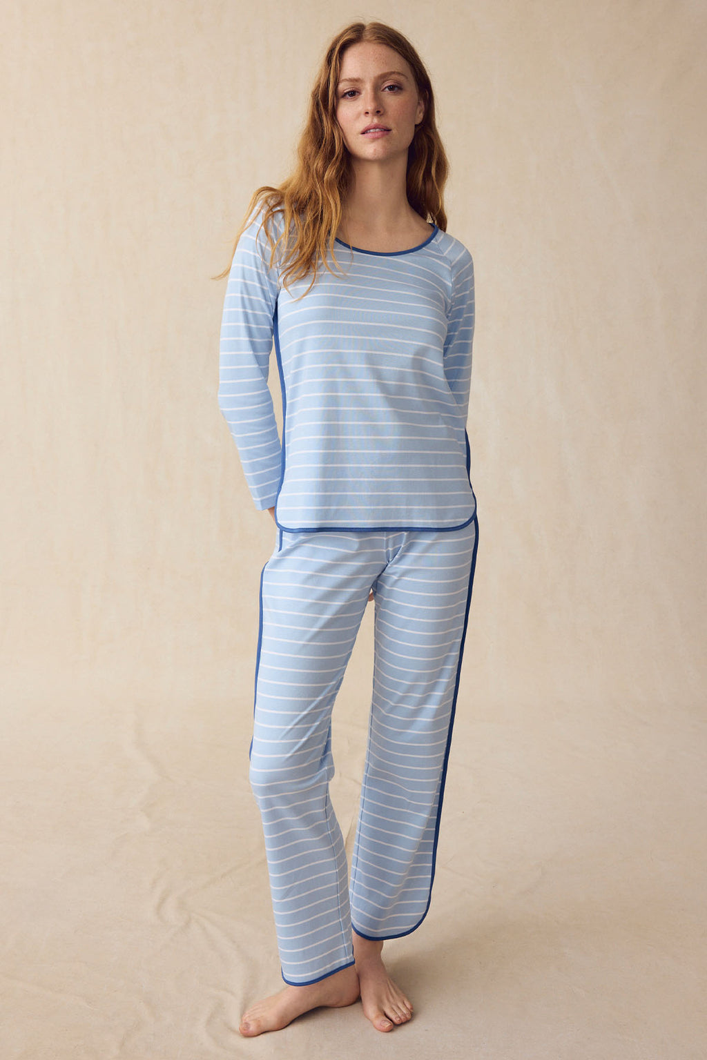 A woman stands on a neutral background wearing the Pima Long Sleeve Bundle in French Blue Club Stripe, crafted from soft Pima cotton with dark blue trim and an elastic waistband.