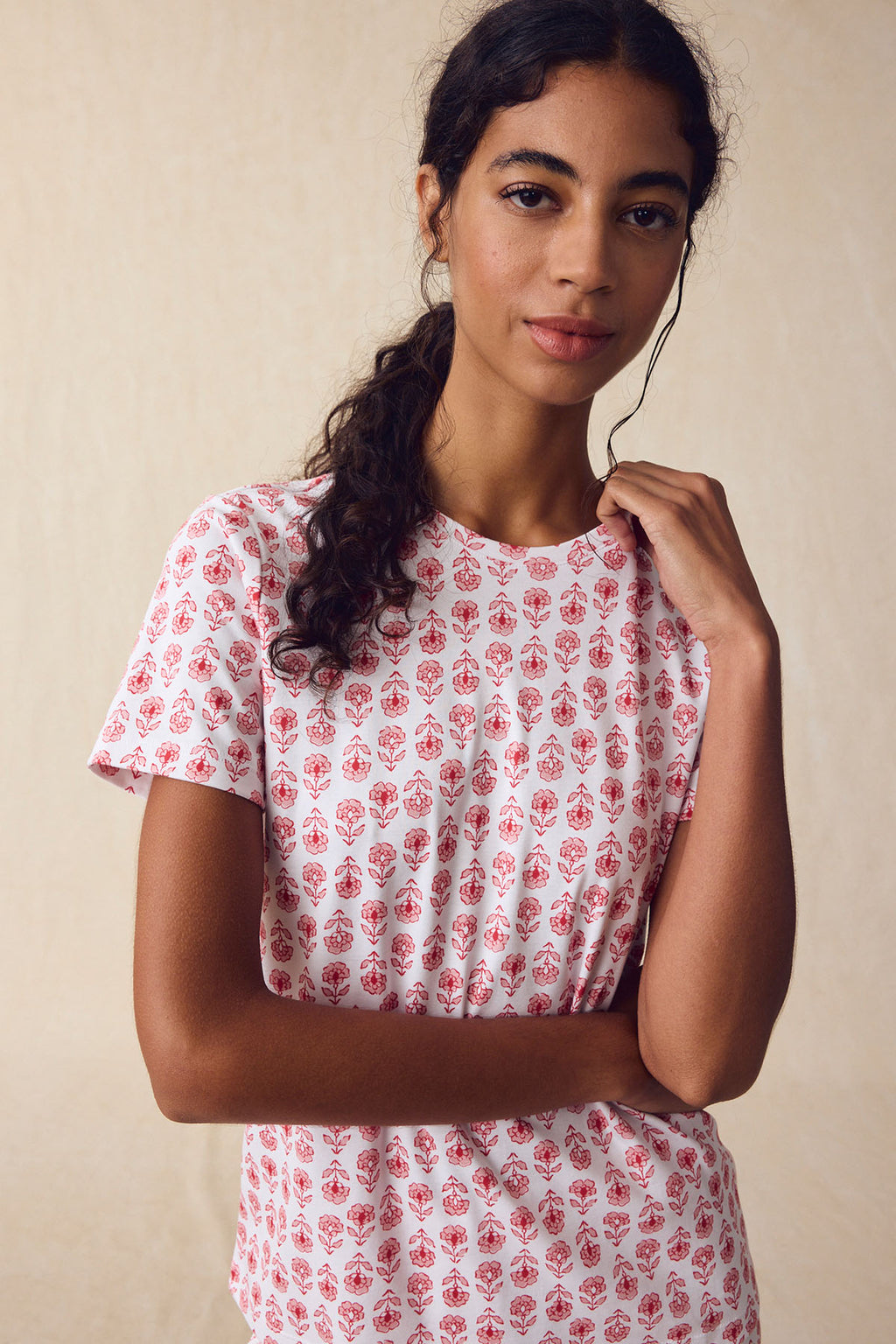 A woman wears the Pima Crew Short-Long Set in Rosewater Sprig, featuring a white Pima cotton T-shirt with a red block print floral pattern, standing against a neutral beige background and looking at the camera.